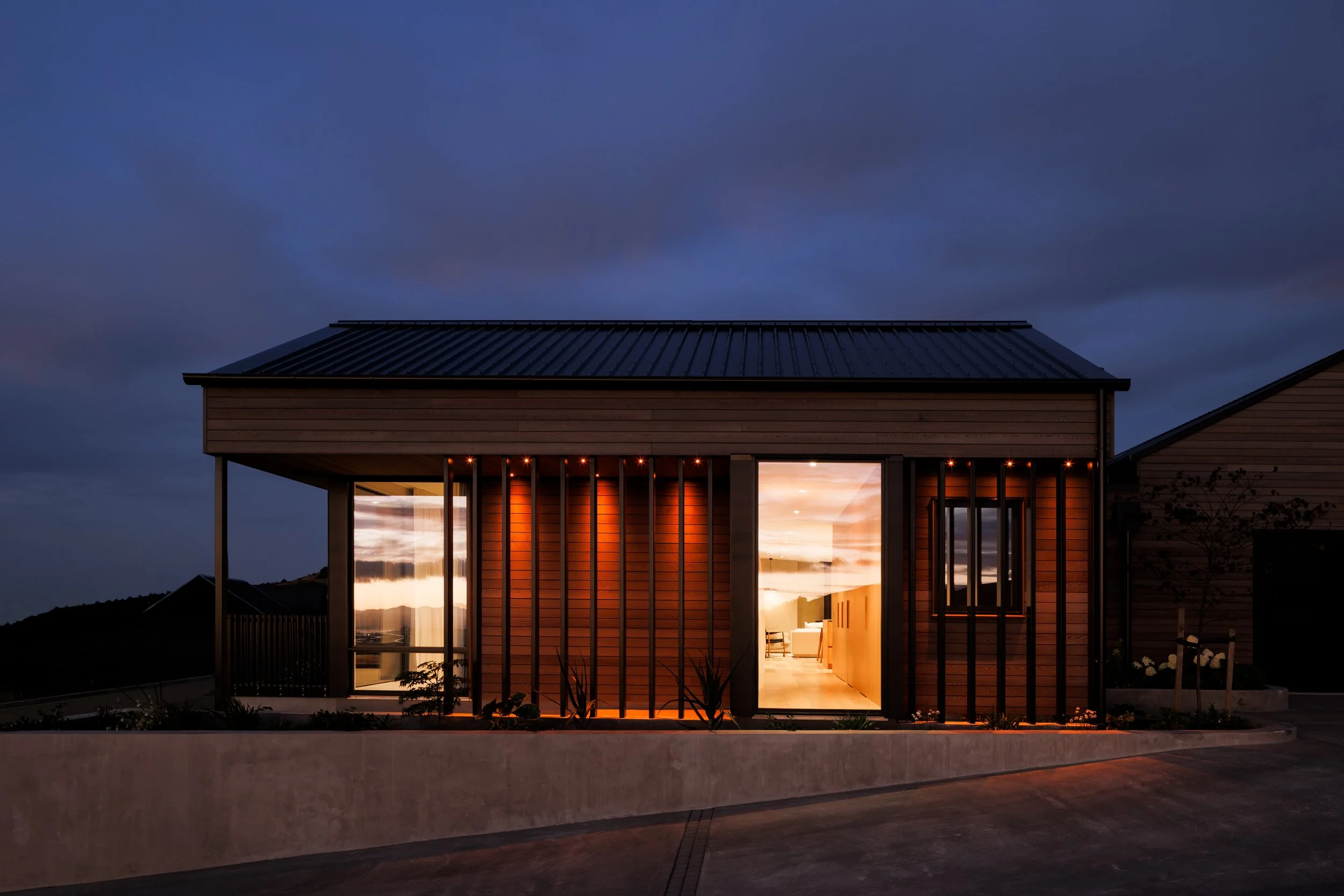 A modern house with large glass windows, illuminated from inside, during twilight, with a cloudy sky in the background.
