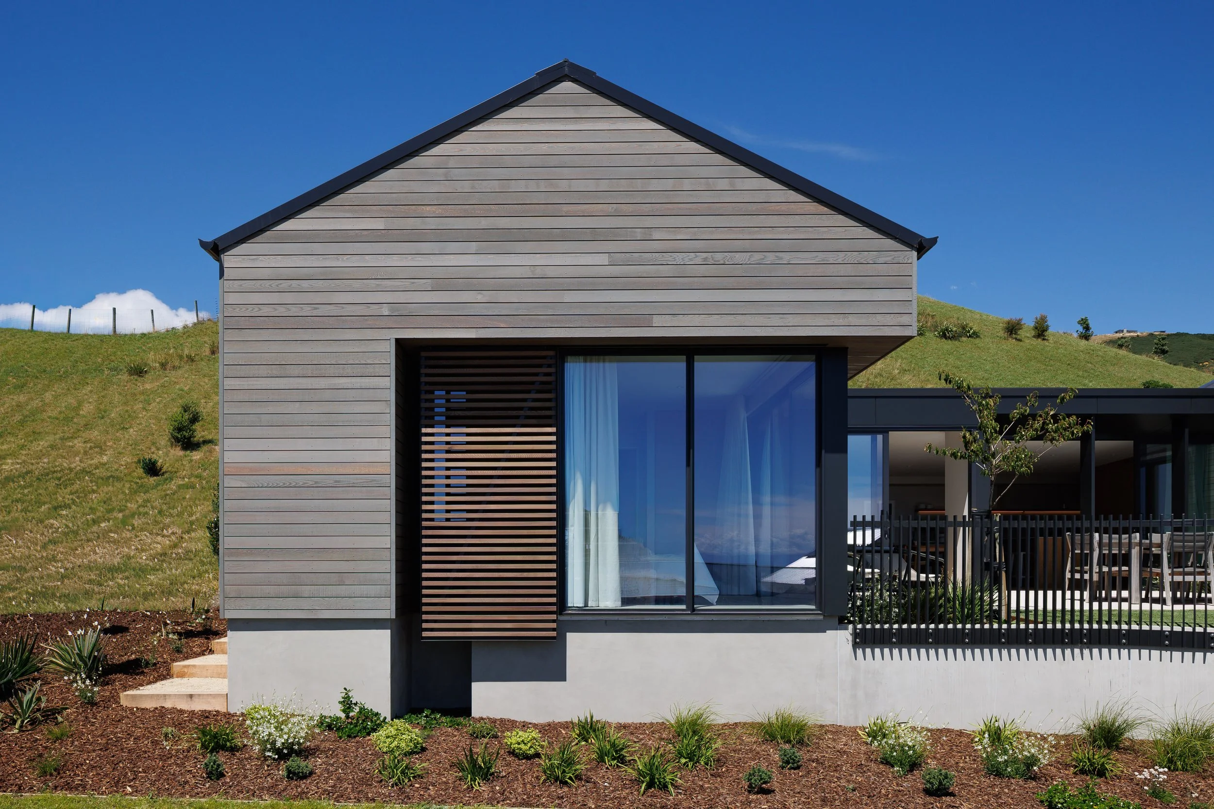 A modern house with a sloped roof, wooden siding, and large glass sliding doors, situated on a grassy yard with small plants and a garden bed in the foreground. The sky above is clear and blue.