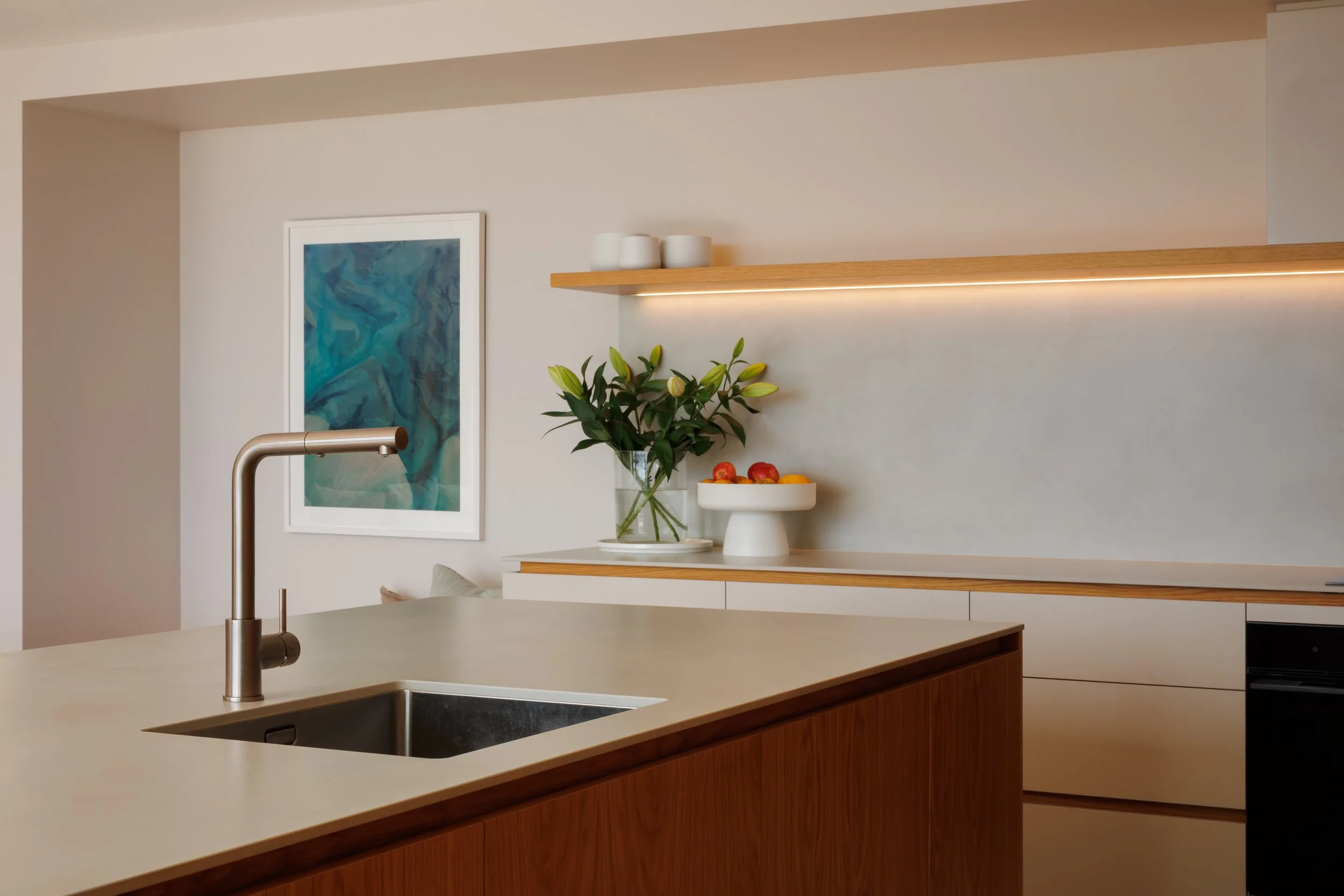 Modern kitchen with a beige countertop island, stainless steel faucet, white cabinetry, a leafy plant in a glass vase, and a bowl of apples and oranges. Artwork hangs on the wall. A wooden shelf with three white cups is above.