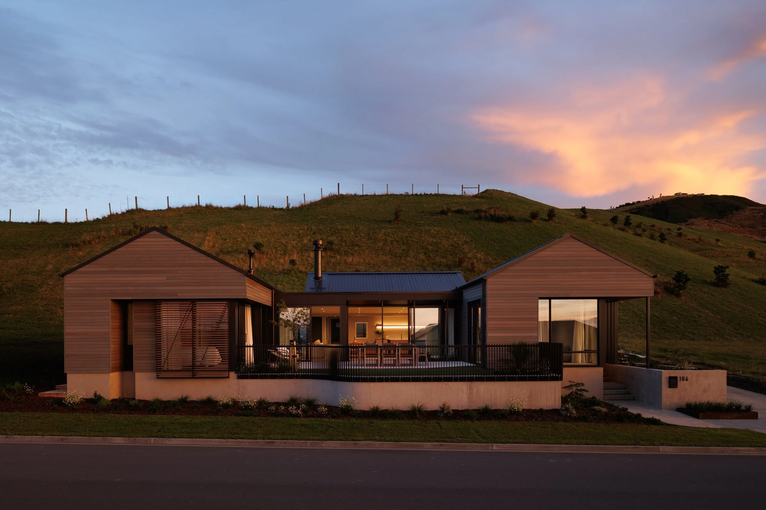 Modern house with large glass windows and wood siding, situated in a hilly area at sunset, with a partly cloudy sky and a fence on the hilltop.
