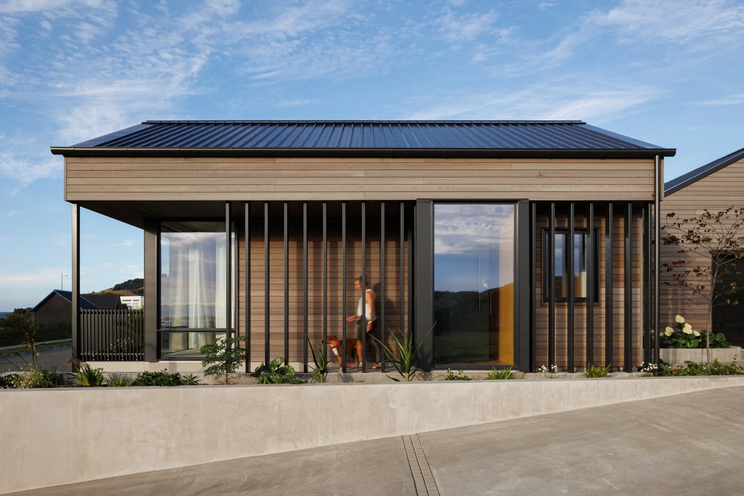 Modern house with wooden siding and large glass windows, situated under a blue sky with wispy clouds. A woman and a dog are seen near the entrance, with a garden and greenery surrounding the house.