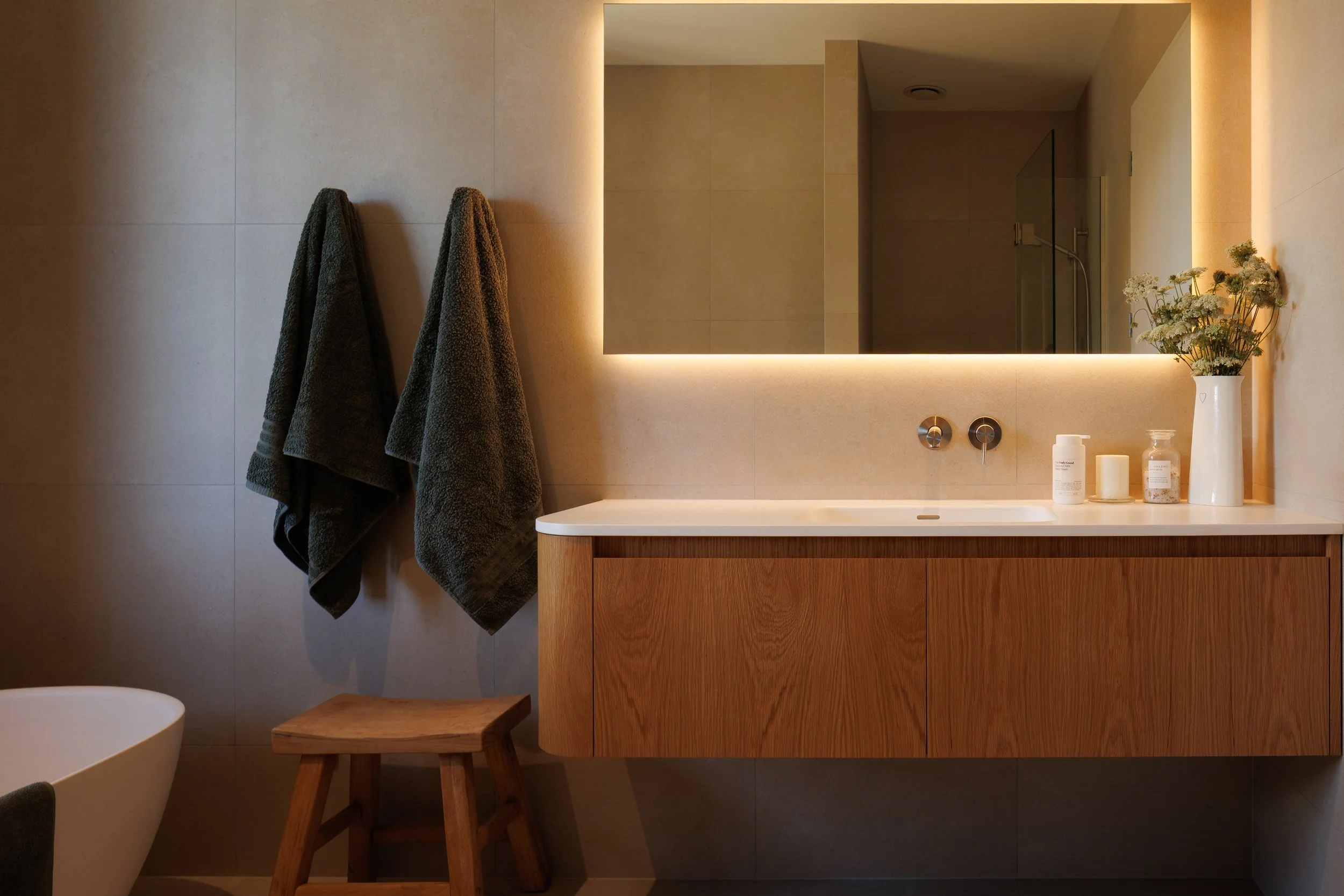 A modern bathroom vanity with a large backlit mirror, beige tiled walls, two dark gray towels hanging on hooks, a white vessel sink, wooden cabinetry, a small wooden stool, and decorative items including a vase with flowers, a soap dispenser, a candl