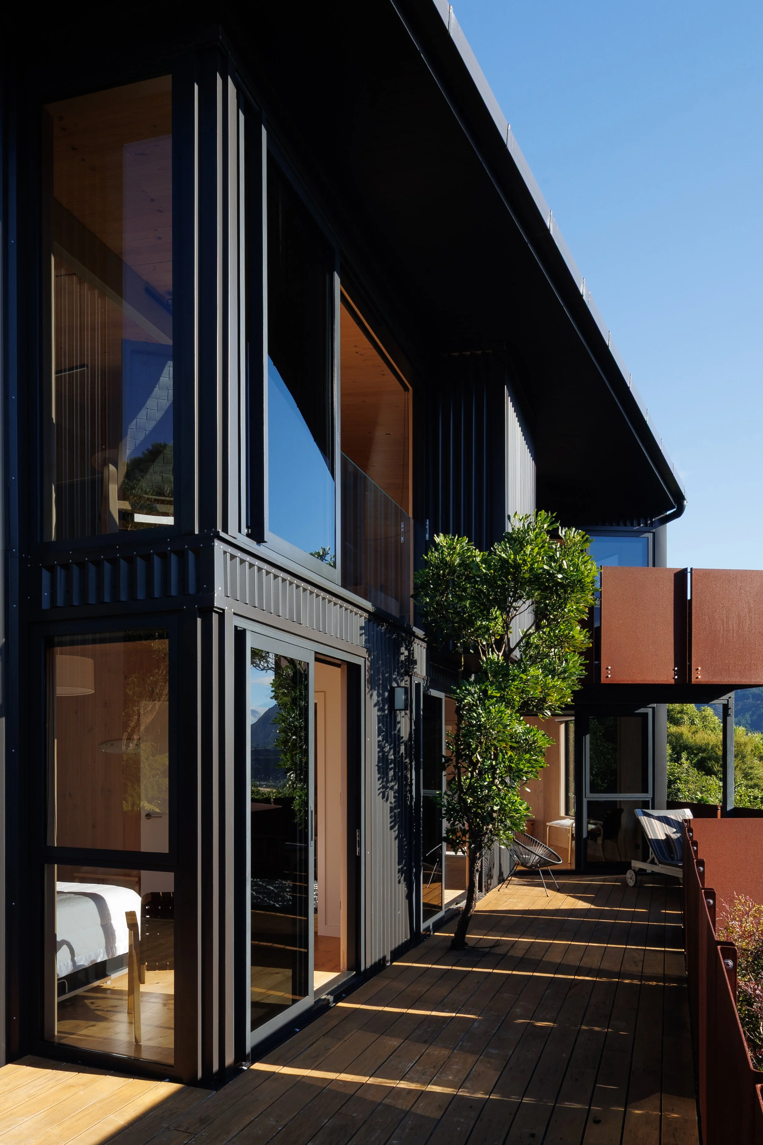 Modern residential building with large glass windows, wooden interior, and an outdoor patio with chairs, a small tree, and a fence under a clear blue sky.