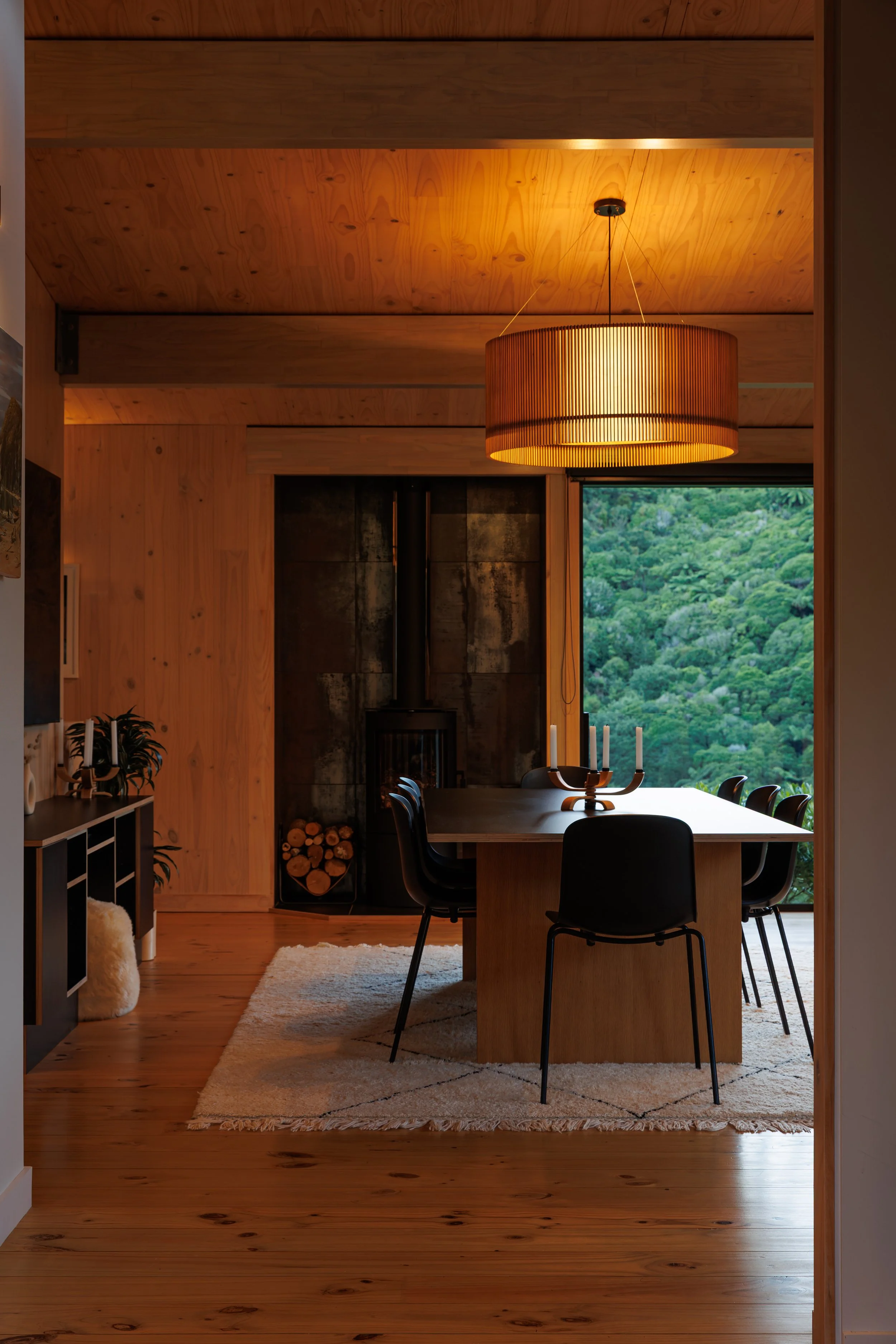 A cozy dining room with wooden walls, ceiling, and floor. A large window shows a green, wooded landscape outside. A round wooden dining table with black chairs is in the center, with a candle holder with four candles. A large round wooden pendant lig