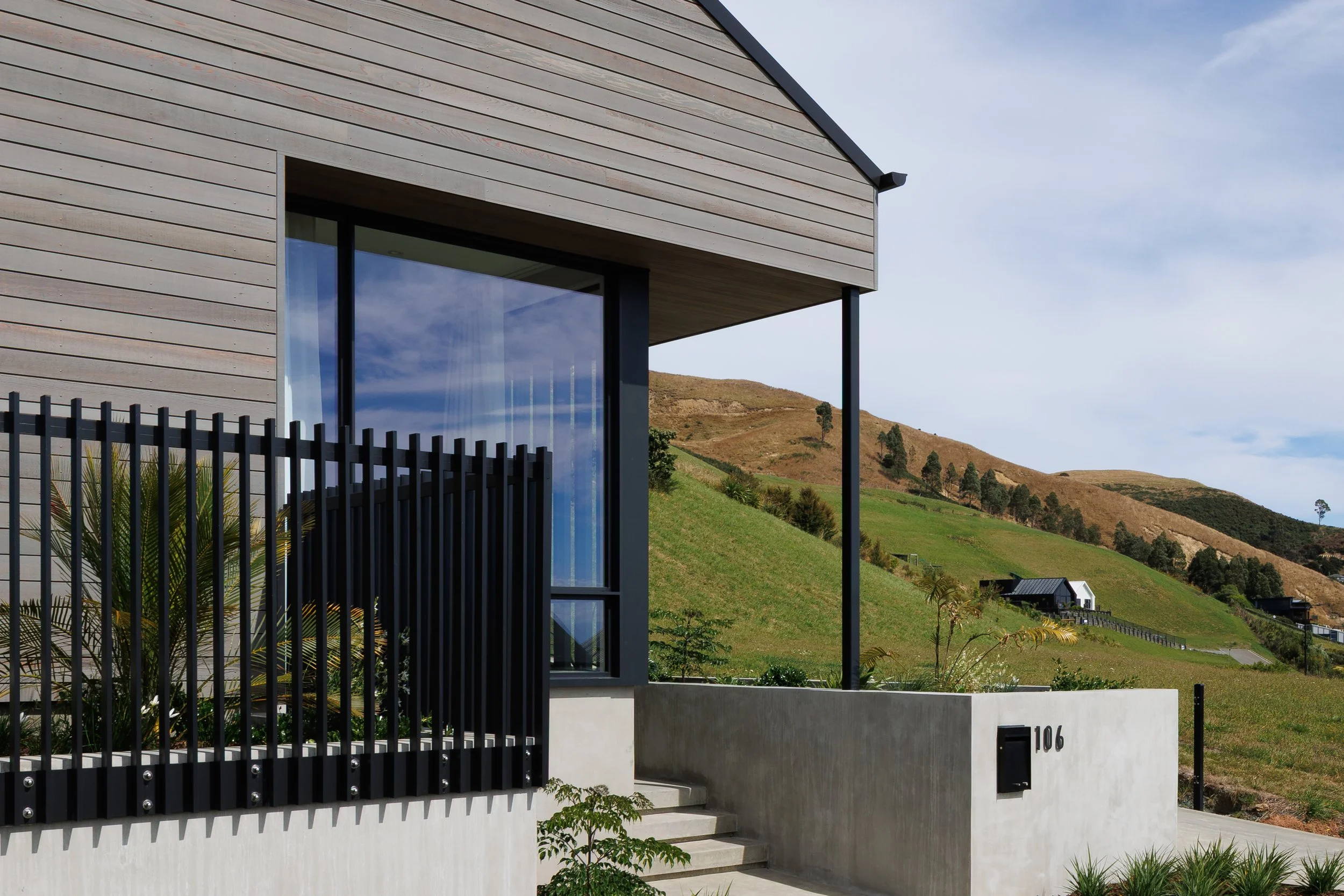 Modern house with large glass window, black metal fencing, and landscape with rolling hills and few trees in background.