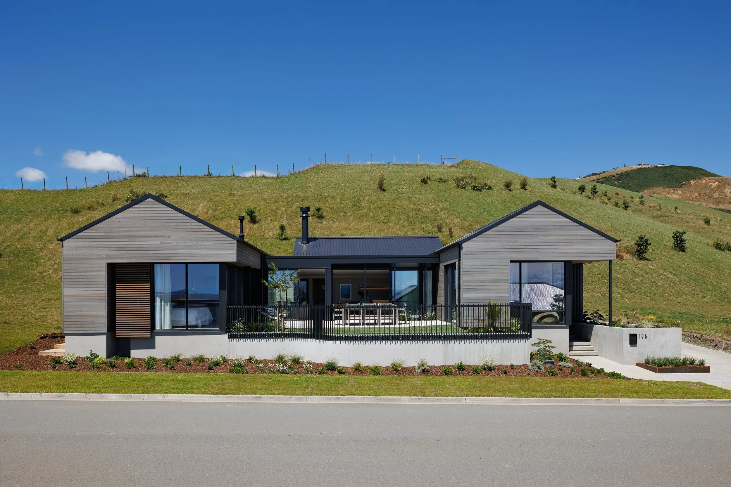 Modern house with gray siding and large glass doors, fenced yard, and landscaped front garden, set against a green hillside under a bright blue sky.