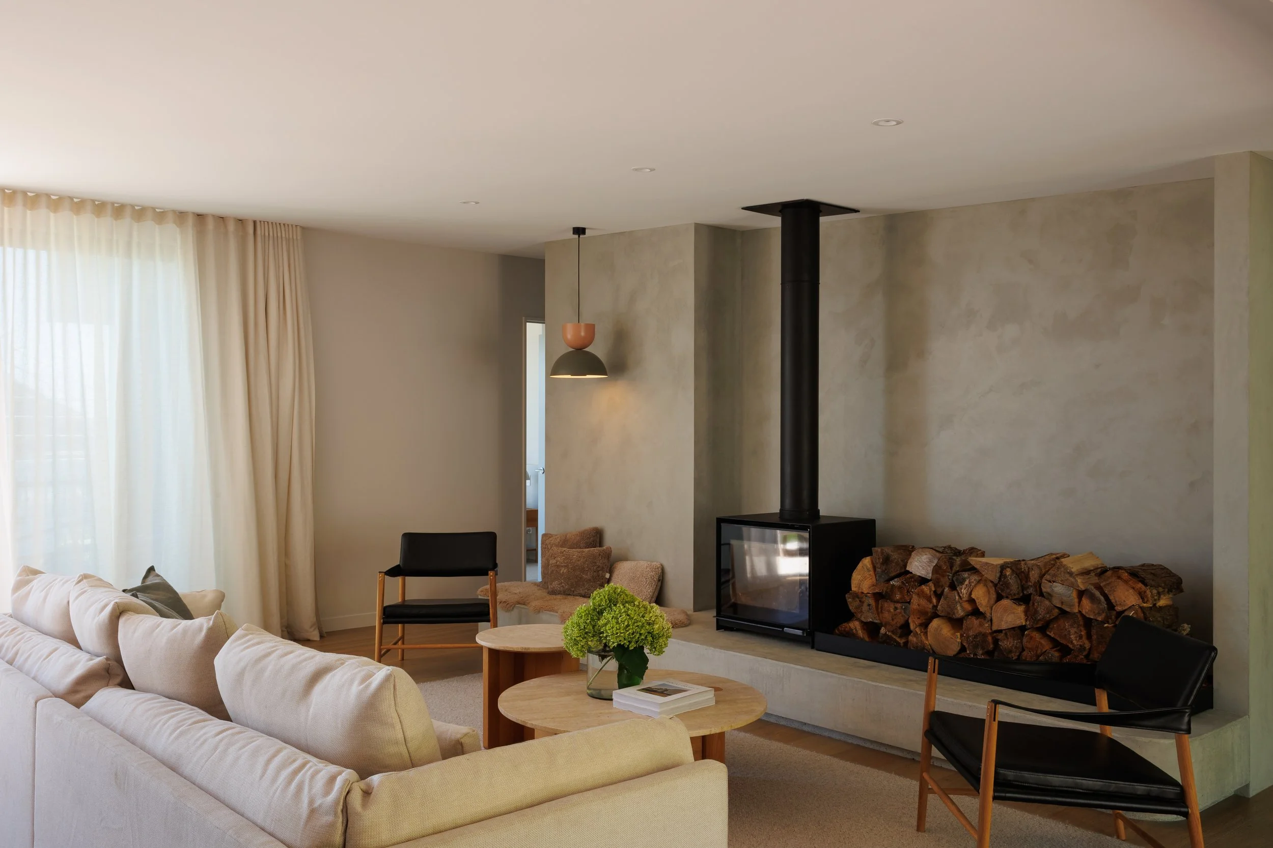 Living room with beige sofa, round wooden coffee table with a green plant and magazines, modern fireplace with stacked firewood, black wood stove, black chairs, cream curtains, and a pendant light.