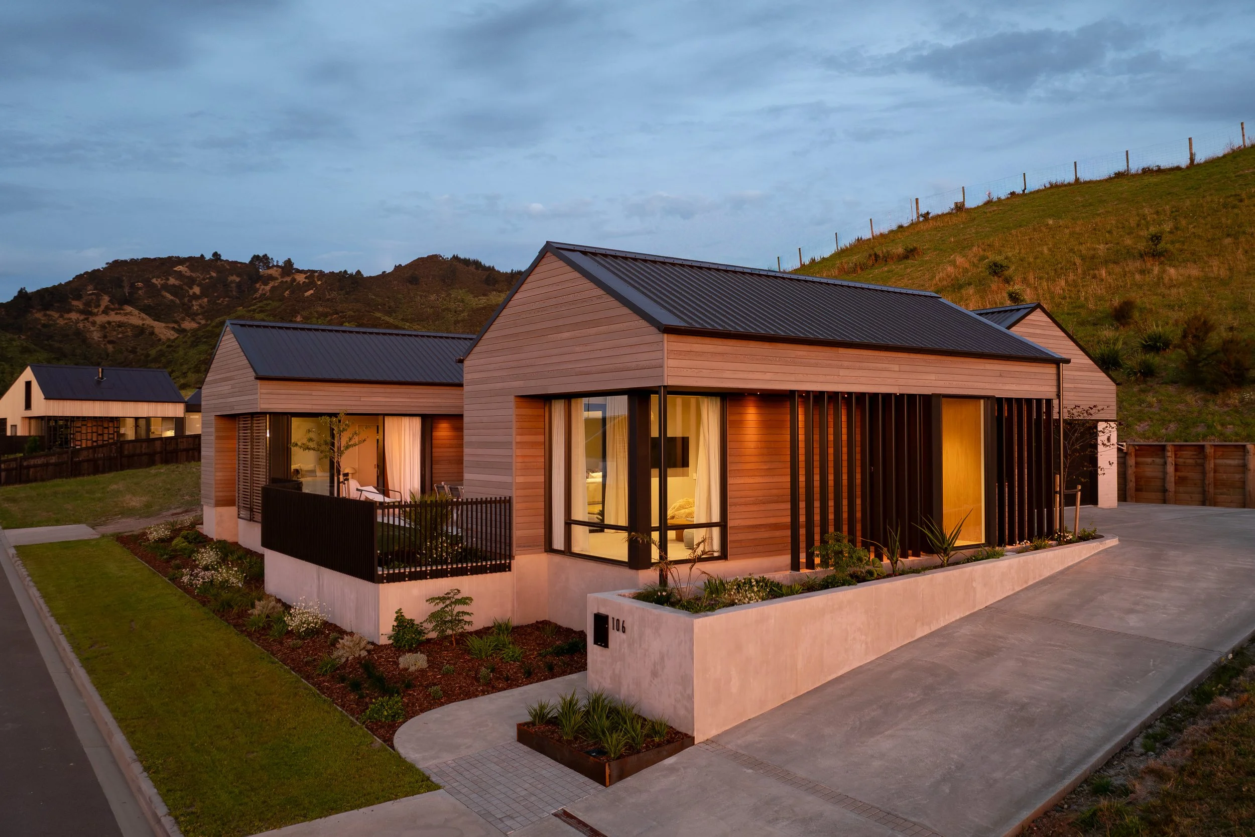Modern house with wooden exterior and large glass windows, situated in a hilly area with green space and landscaping, during dusk.