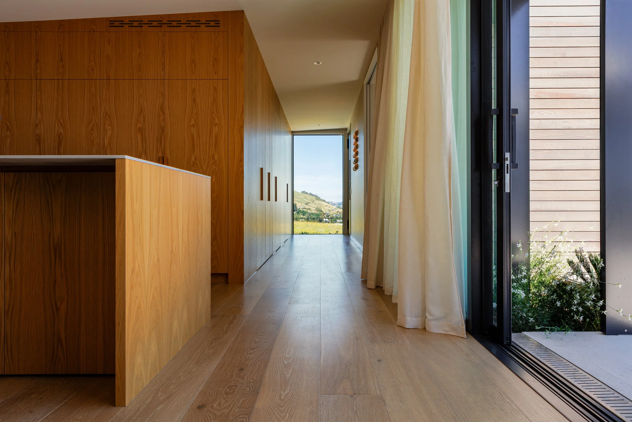 Interior of a modern house with wooden walls and floor, large glass door with curtains, and a view of a hilly landscape outside.