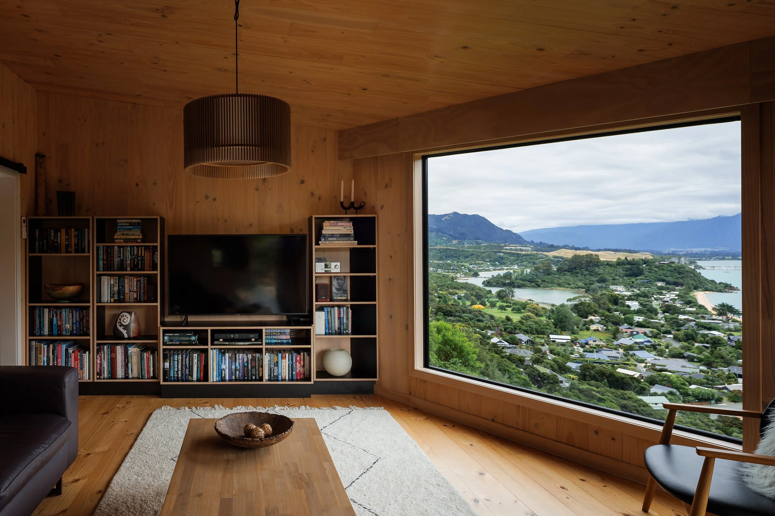 Living room with wooden walls and ceiling, large window overlooking a scenic landscape of mountains, water, and houses, with a bookshelf, TV, and a table with decorative items.