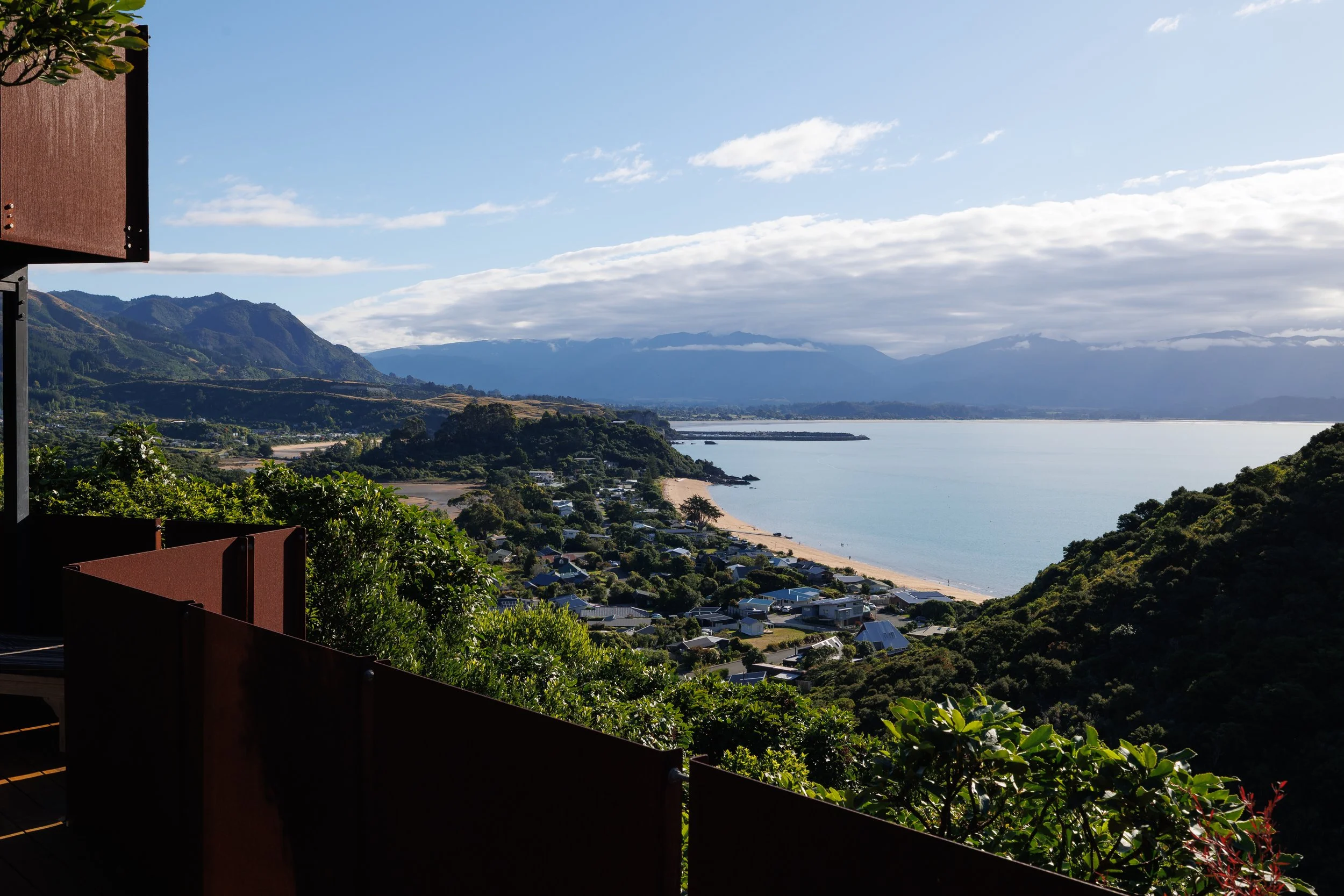 Scenic view of a coastal landscape with houses, a beach, water, and mountains in the background, seen from a balcony with greenery.