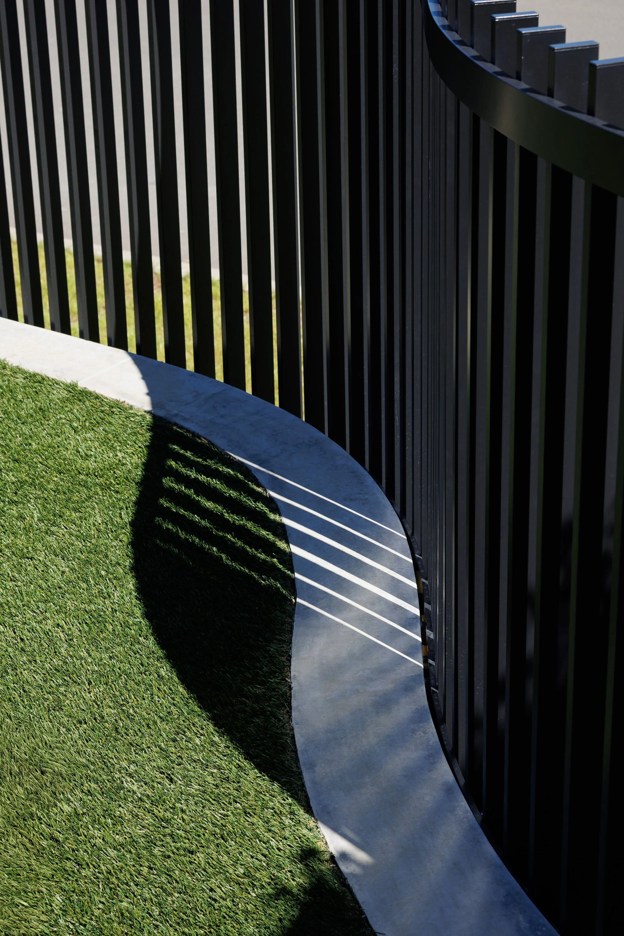 Close-up view of a black metal fence with vertical bars casting shadows on a curved concrete pathway and green grass below.