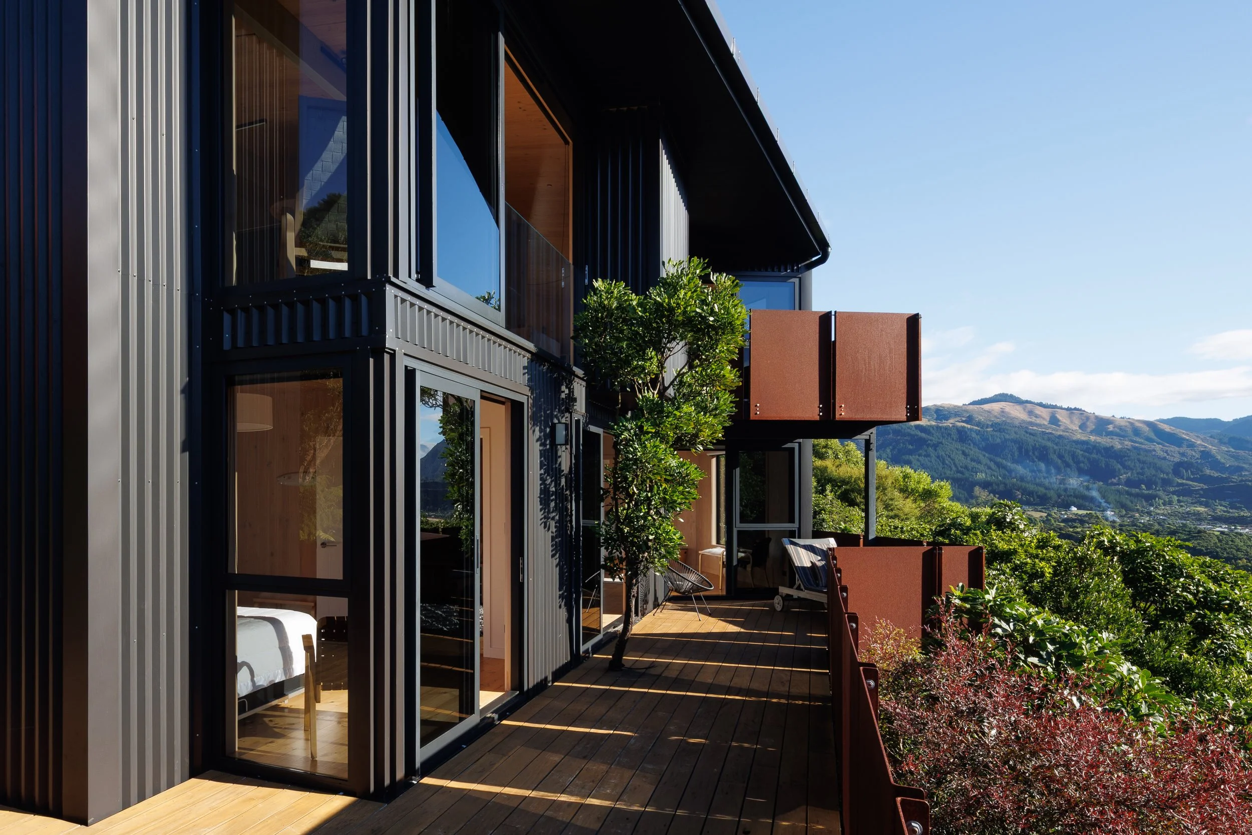 Modern multi-story building with large glass windows and a balcony, surrounded by greenery and mountains in the background, under a clear blue sky.
