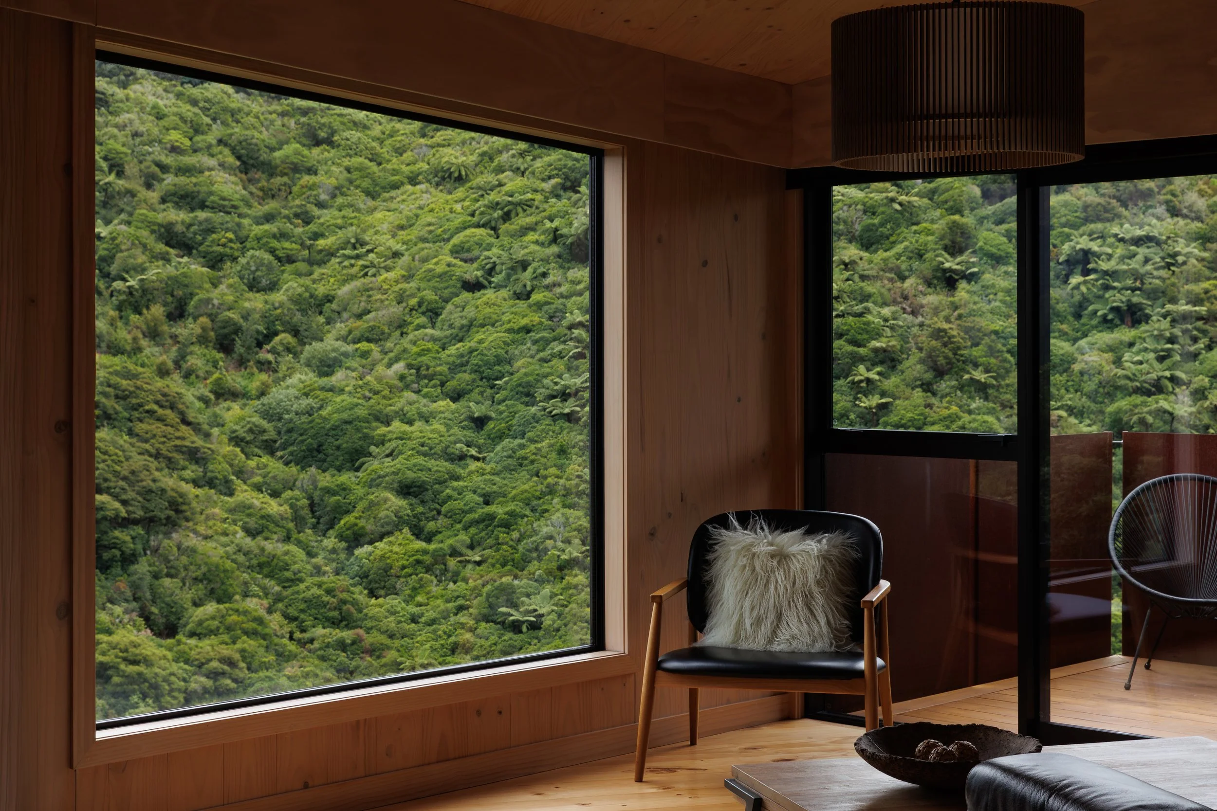 A cozy interior room with large windows showing lush green forest outside. The room has wooden walls and flooring, a black chair with a furry pillow, and a modern hanging ceiling light.