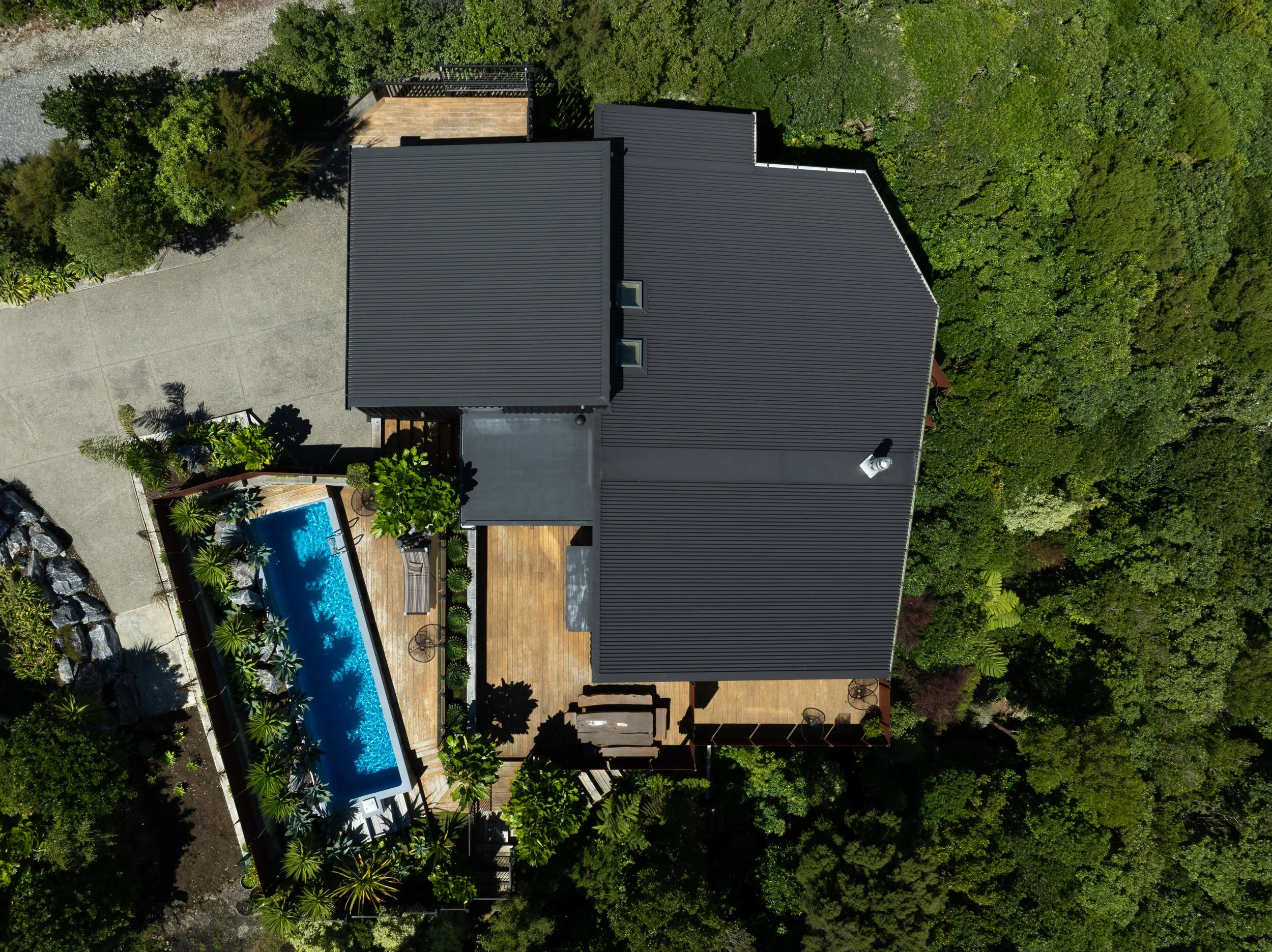 Aerial view of a house with a black metal roof, a swimming pool, a wooden deck area, surrounded by green trees and plants.