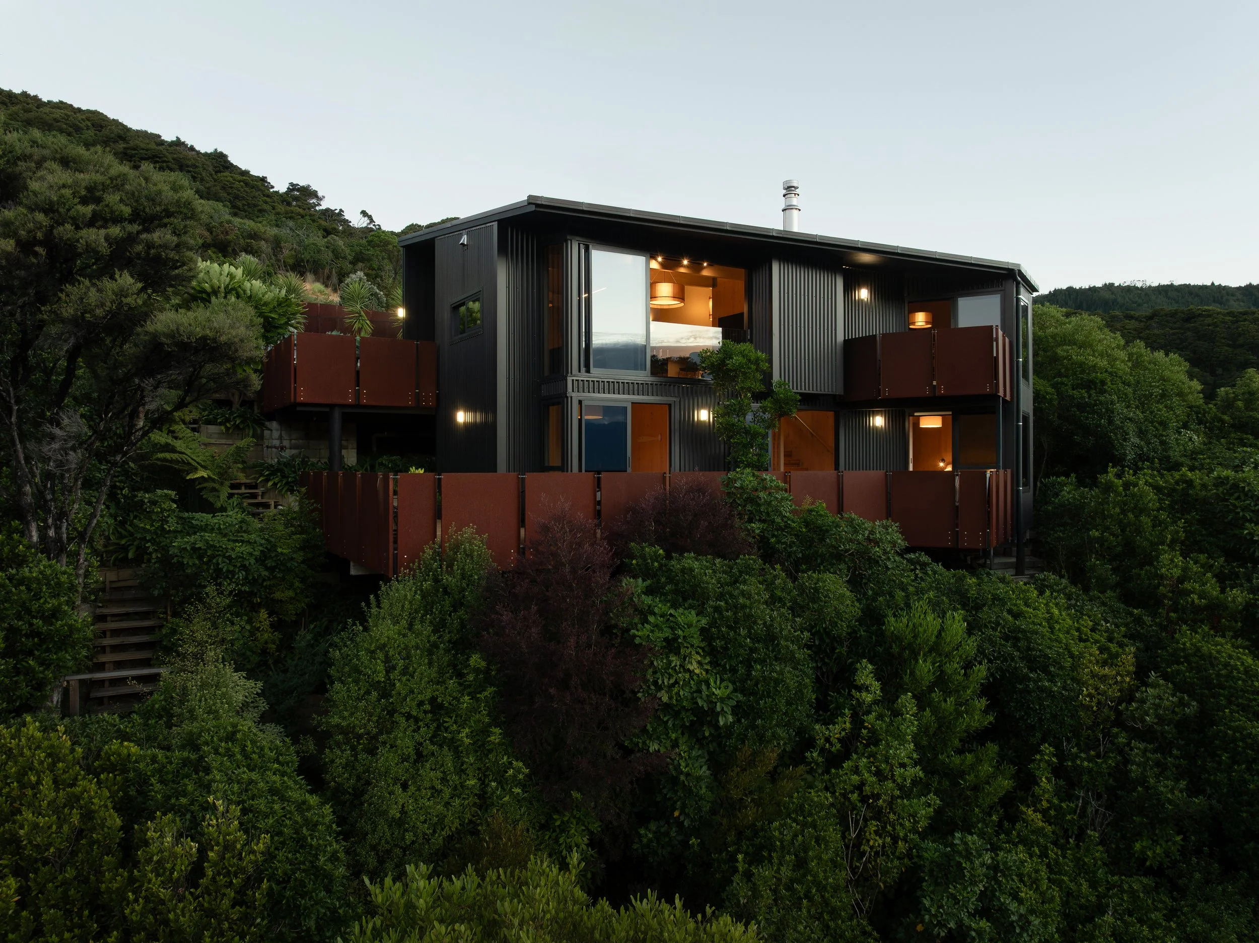 A modern black house with large glass windows and a balcony, nestled among green trees and bushes on a hillside, with a clear sky in the background.
