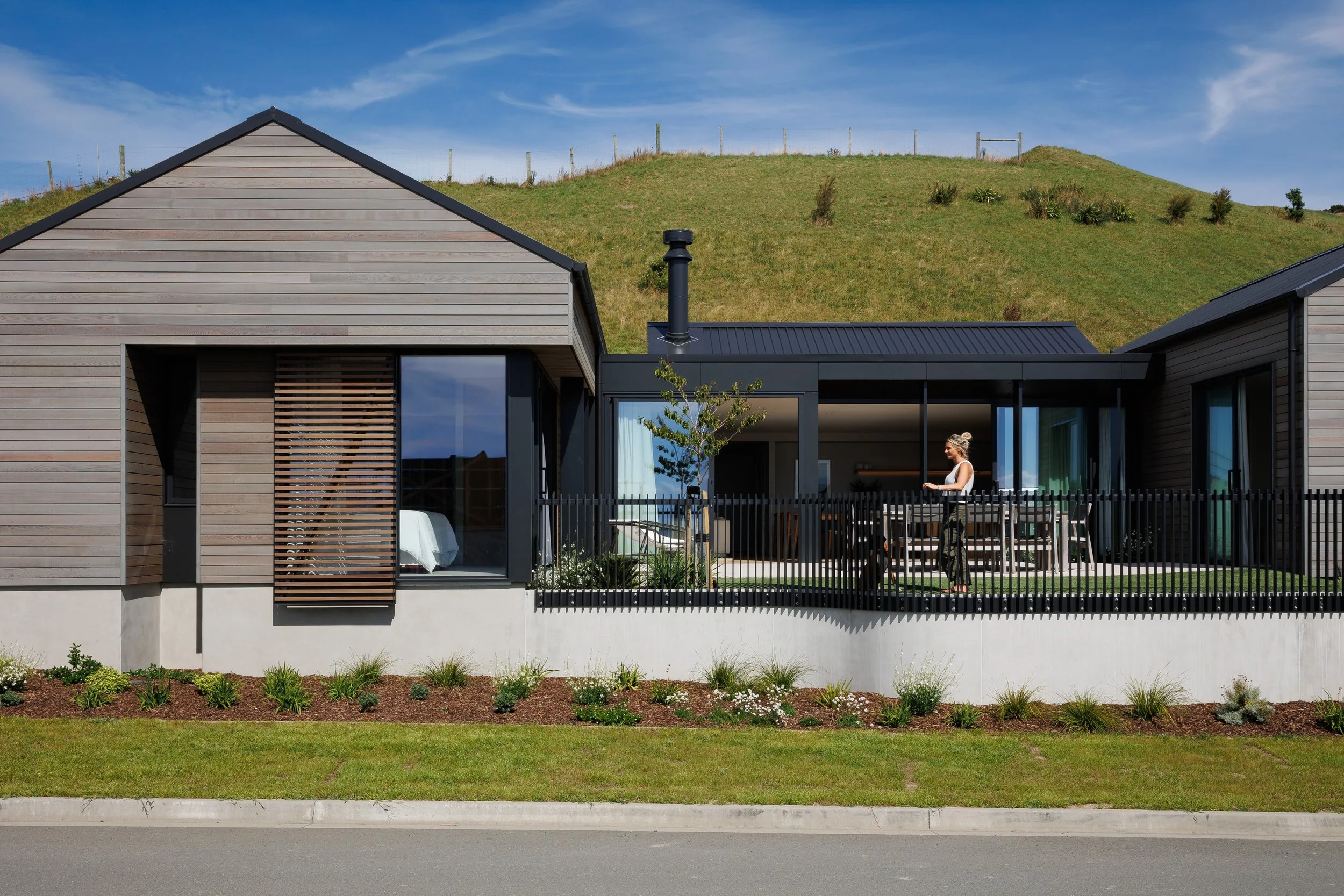 Modern house with wooden siding, large windows, a backyard patio with outdoor furniture, a garden with plants, and a green hill in the background under a blue sky.