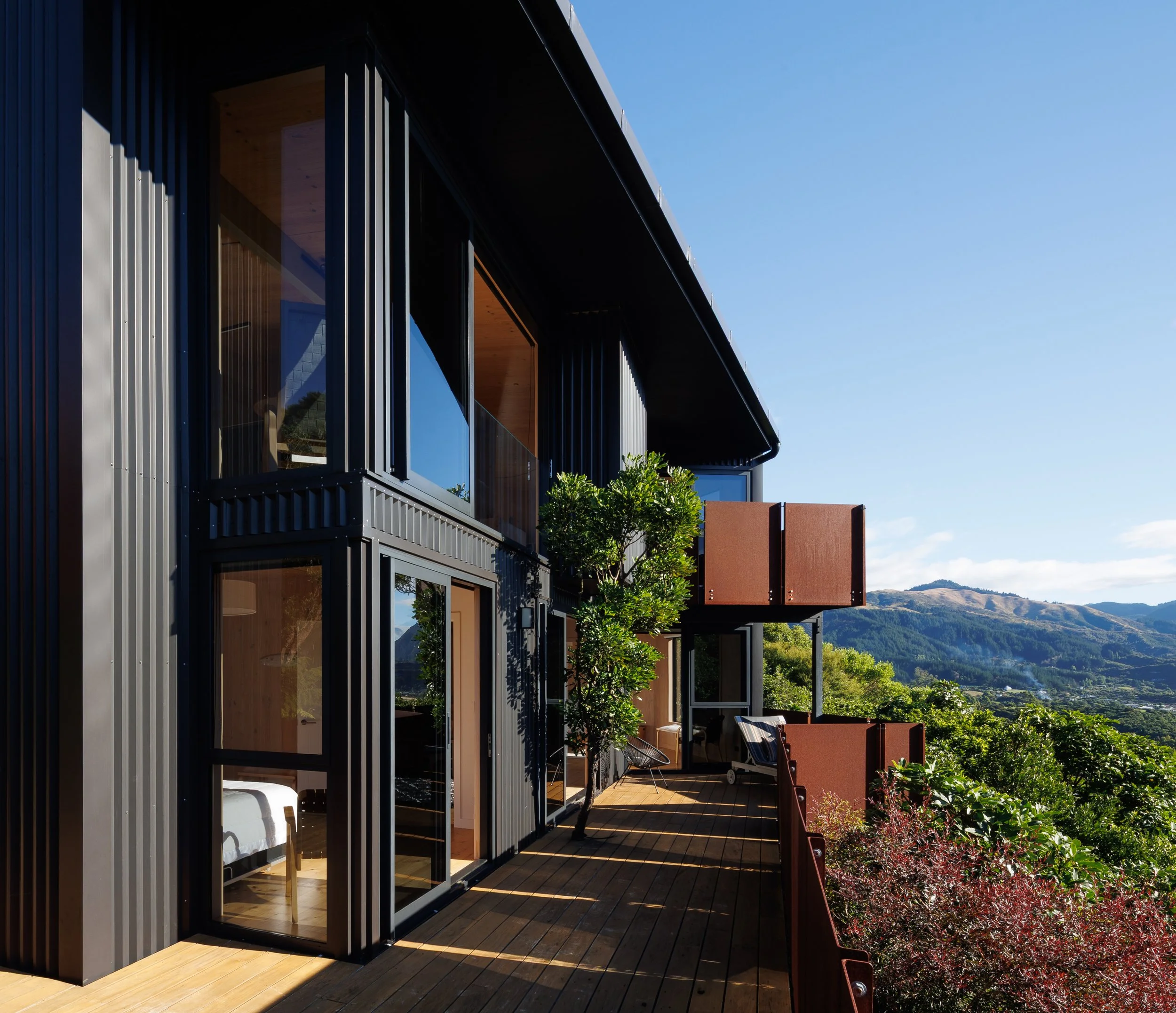 Modern multi-story house with large glass windows and wooden and metal siding, overlooking a lush green landscape with mountains in the background, under a clear blue sky.