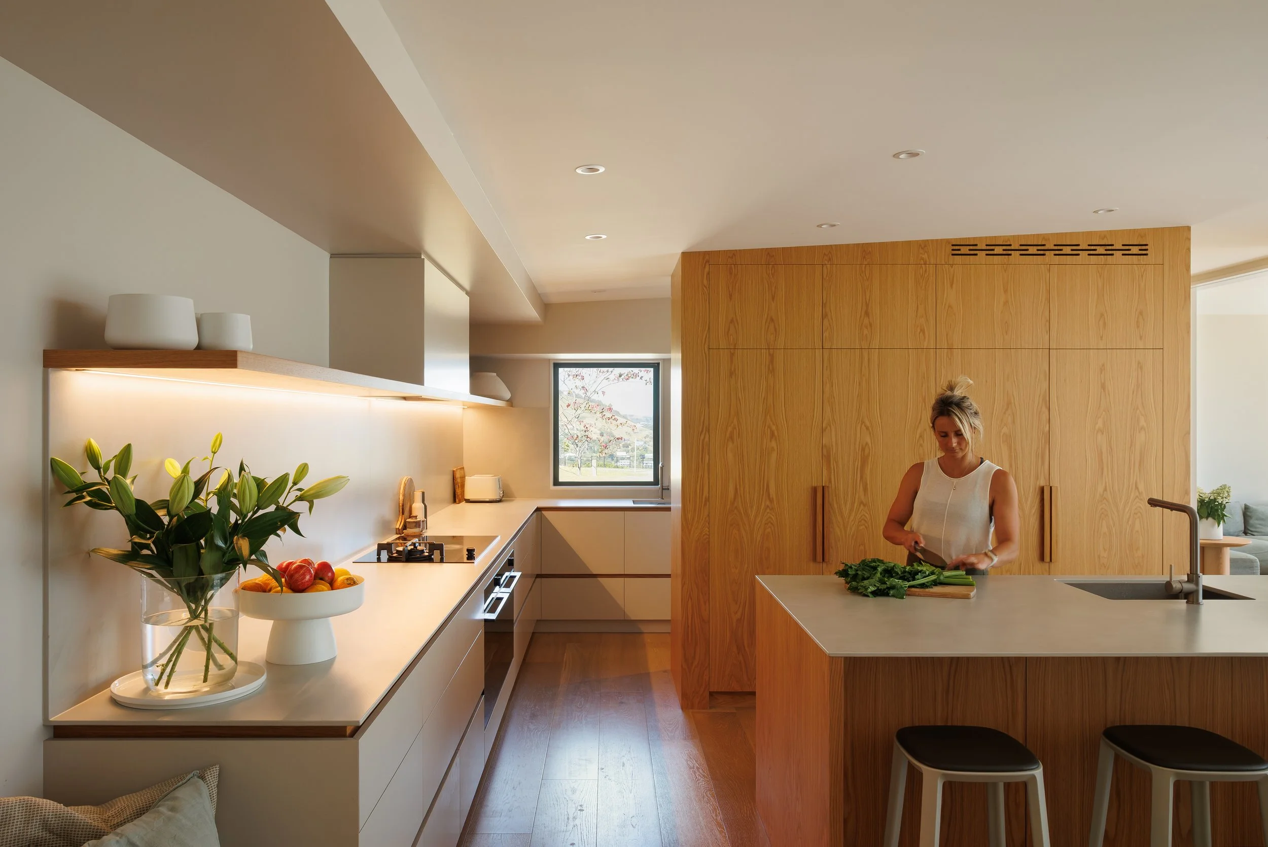 A woman chopping greens at a kitchen island with a window in the background showing a tree. The kitchen features wooden panels, white cabinets, and a countertop with a bowl of fruit and a vase of flowers.