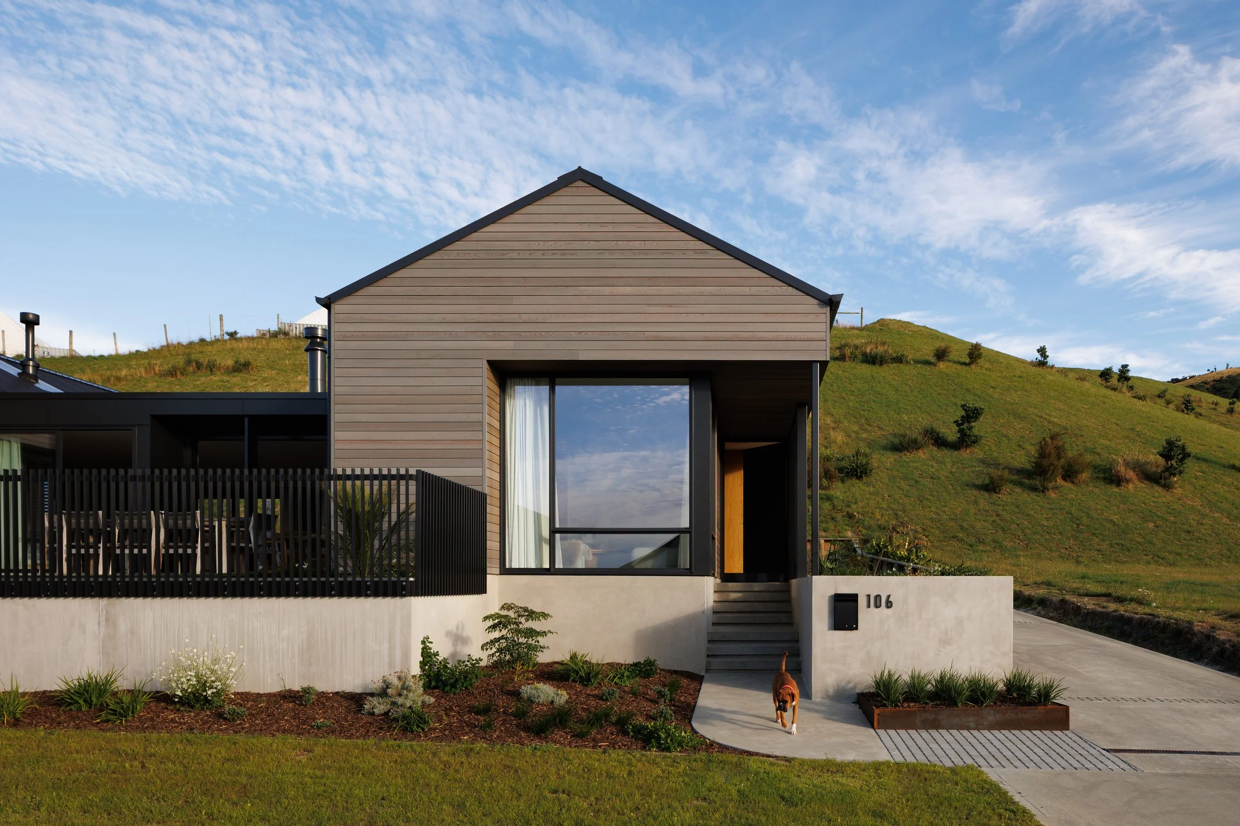 Modern house with a sloped roof, large glass window, and a small front porch, situated on a grassy hillside under a partly cloudy sky, with a small dog near the entrance.