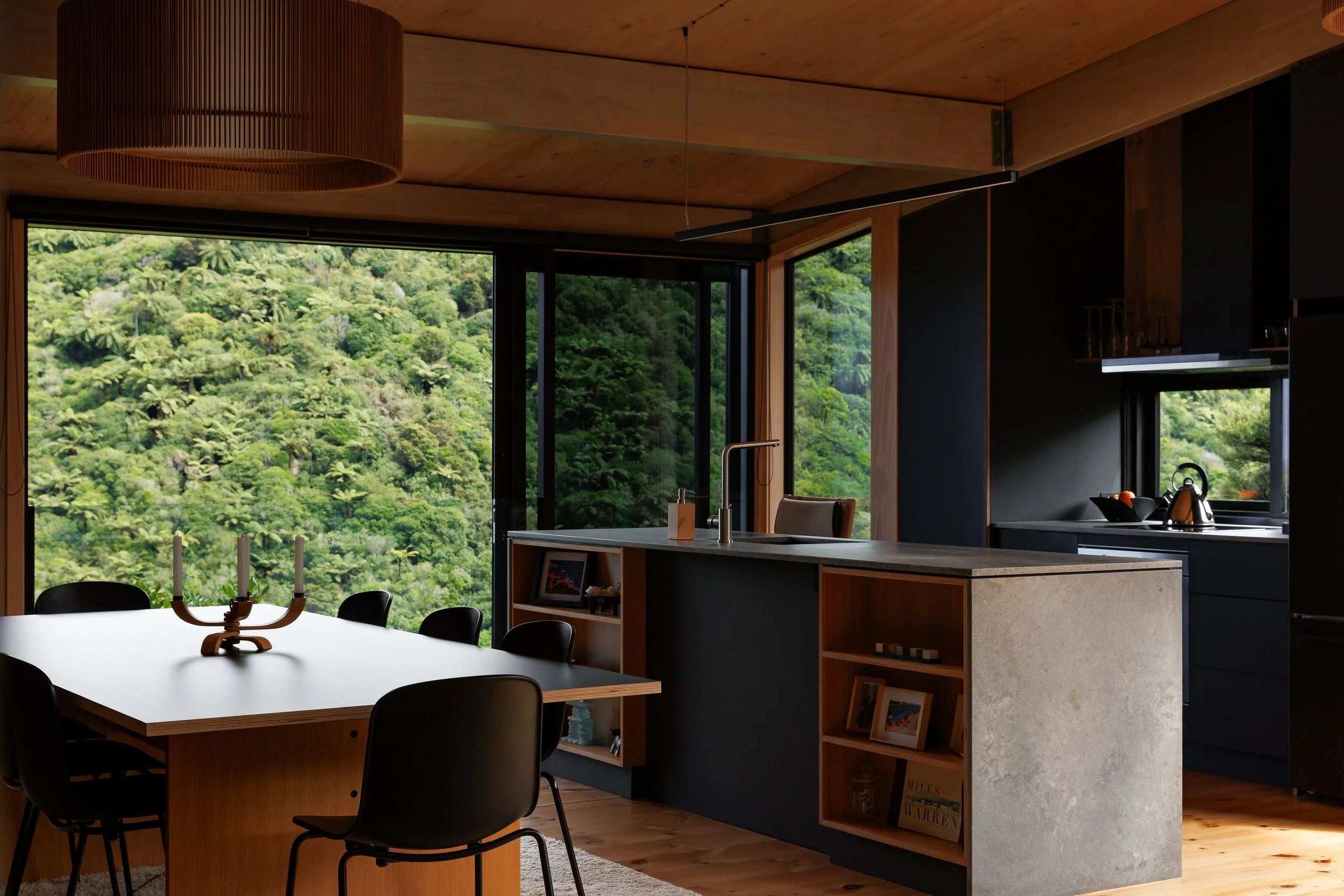 Modern dining area with a view of lush green forest outside large windows, a white dining table with black chairs, and a kitchen with dark cabinetry and a kitchen island.