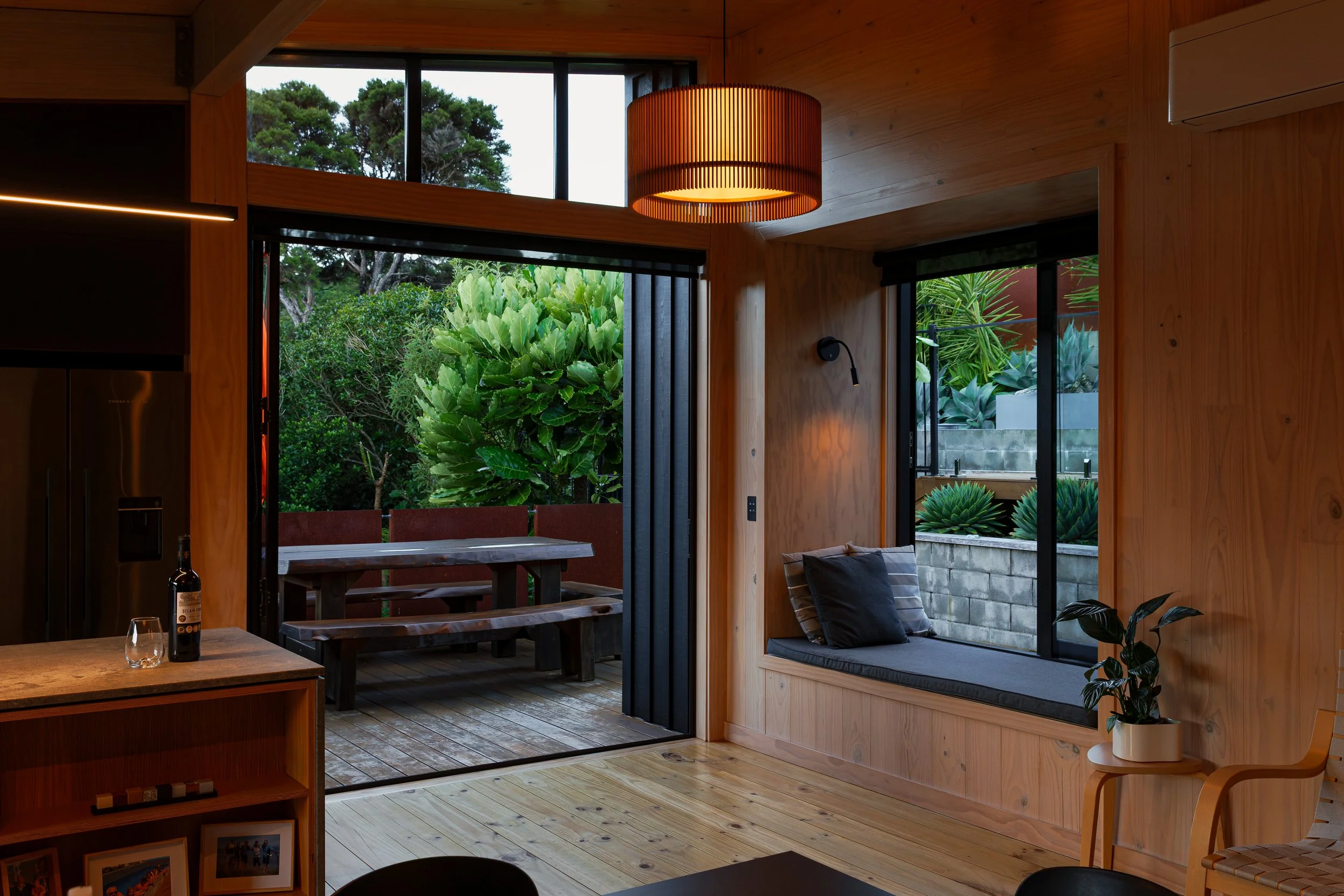 Interior of a modern wooden house with large glass sliding doors opening to a garden with lush green plants and trees. The room features a wooden ceiling, a hanging lamp, and cozy seating area with cushions. There are also bottles and glasses on a wo