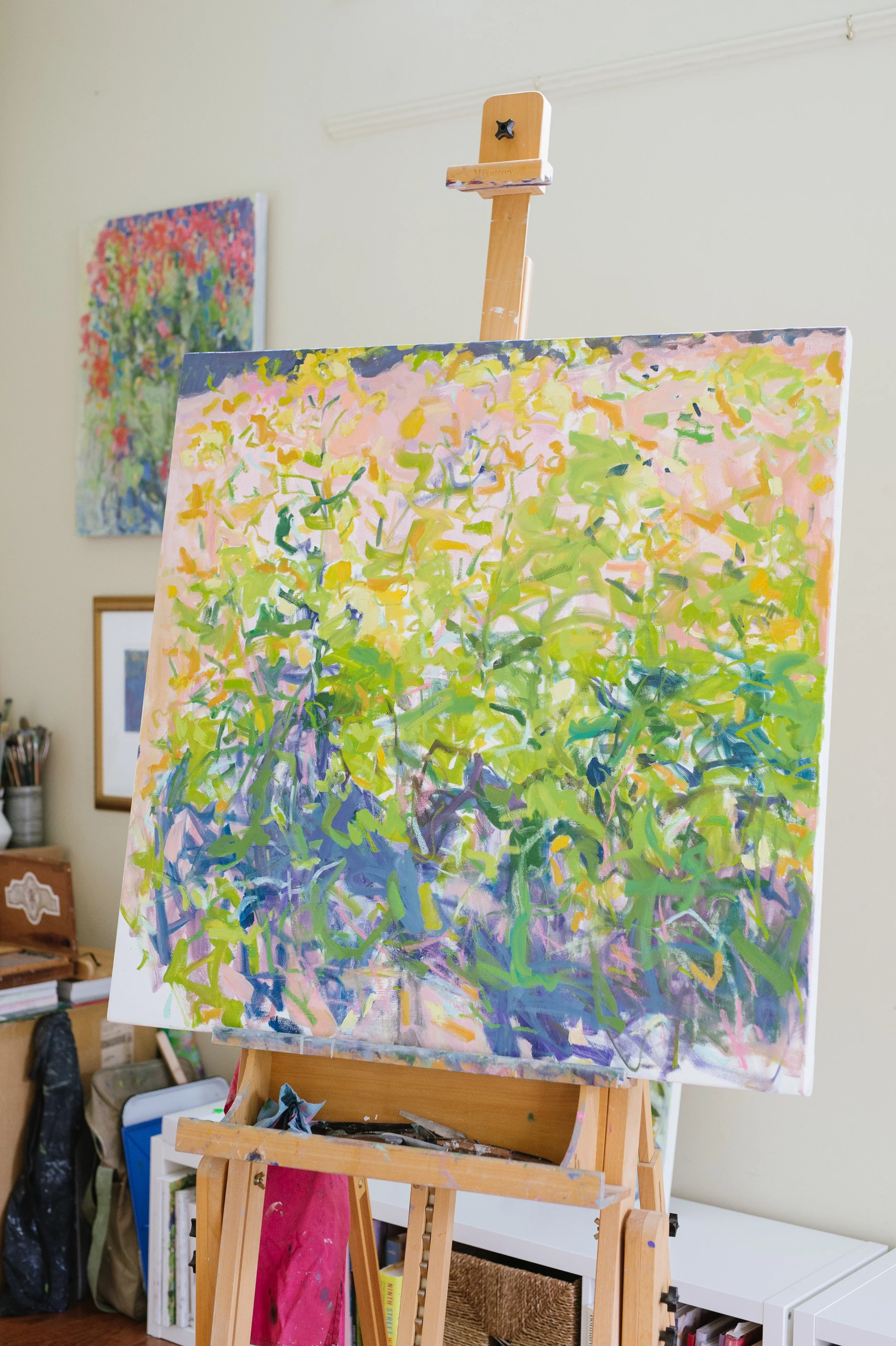 An abstract expressionist painting on an easel, with vibrant colors of pink, yellow, green, and purple, in an art studio. Two paintings are visible on the wall in the background.