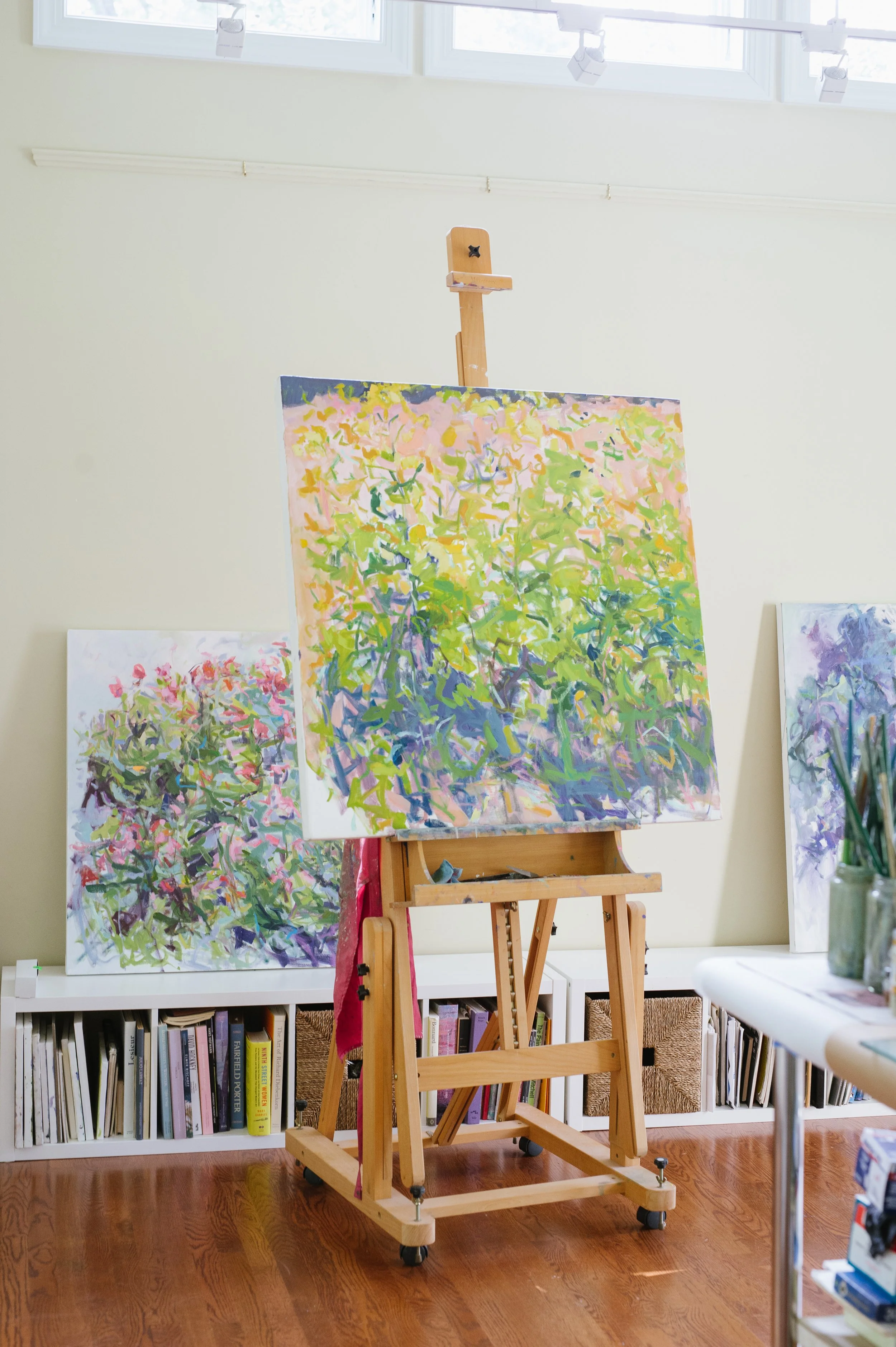 An art studio with a blank wooden easel on wheels in front of a wall. Behind the easel, there are three colorful abstract paintings leaning against the wall, with bookshelves below filled with various books. A window with bright, natural light illumi