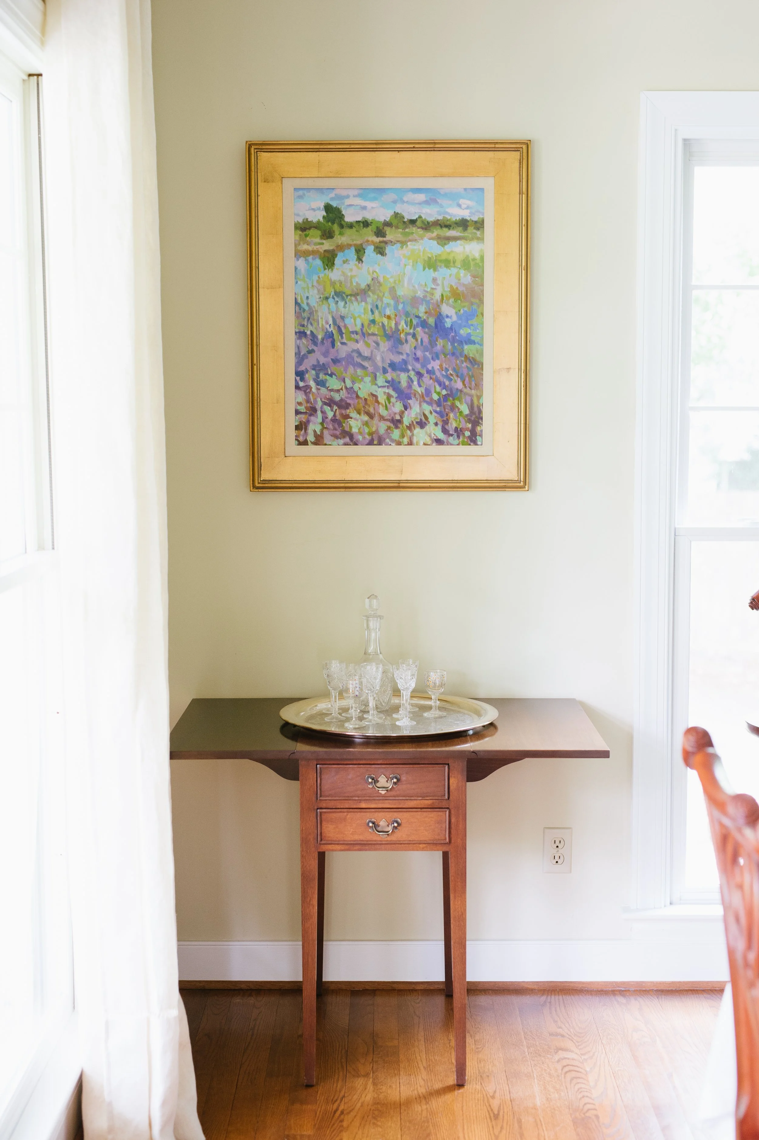 A wooden side table with drawers holding a silver tray with glassware, positioned beneath a framed landscape painting and near a window with white trim in a room with light-colored walls and hardwood floors.