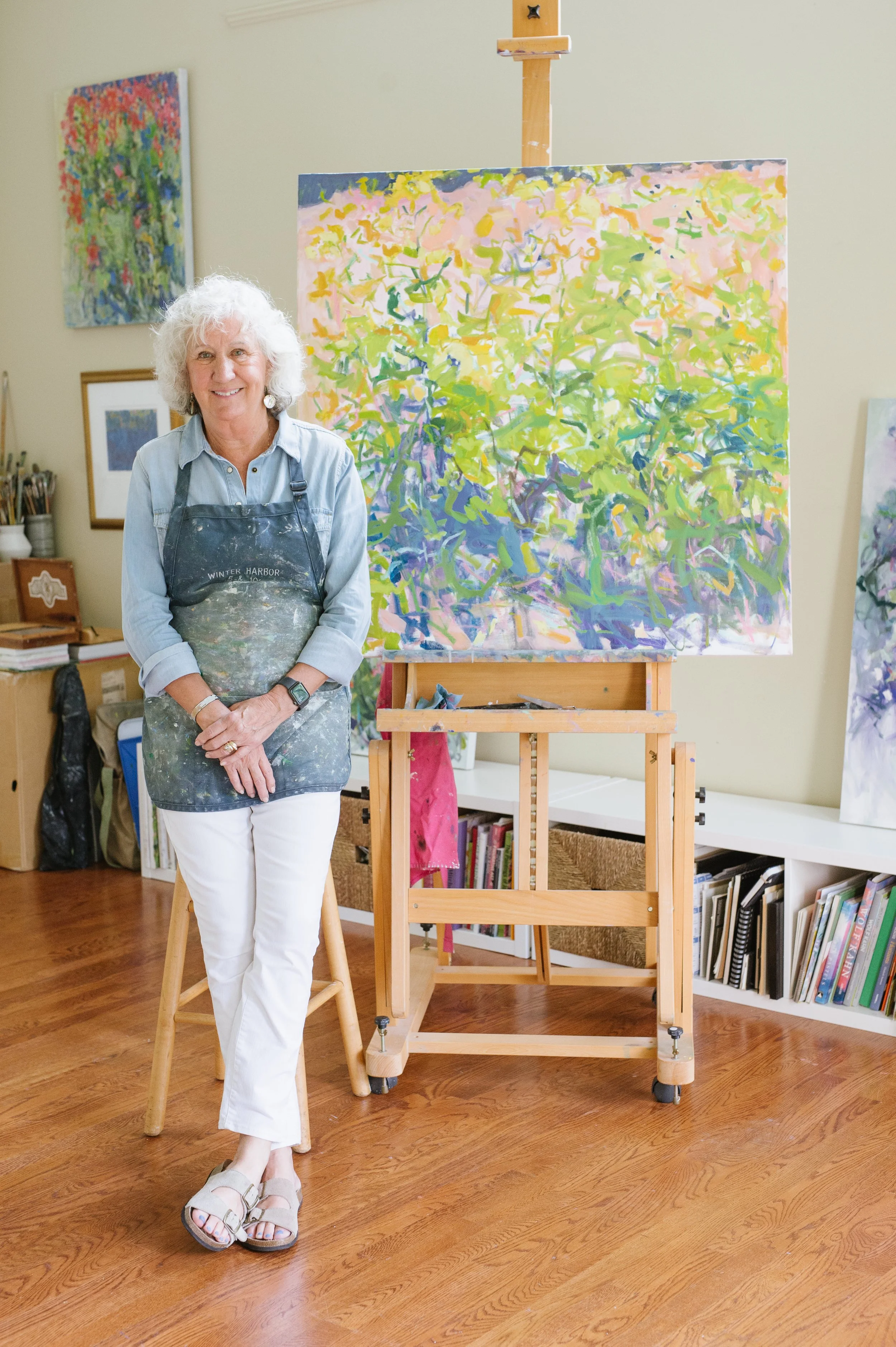 Priscilla Whitlock smiling and standing next to her easel with a colorful abstract painting on it in an art studio. She is wearing a denim shirt, white pants, and sandals, and has an apron with paint splatters.