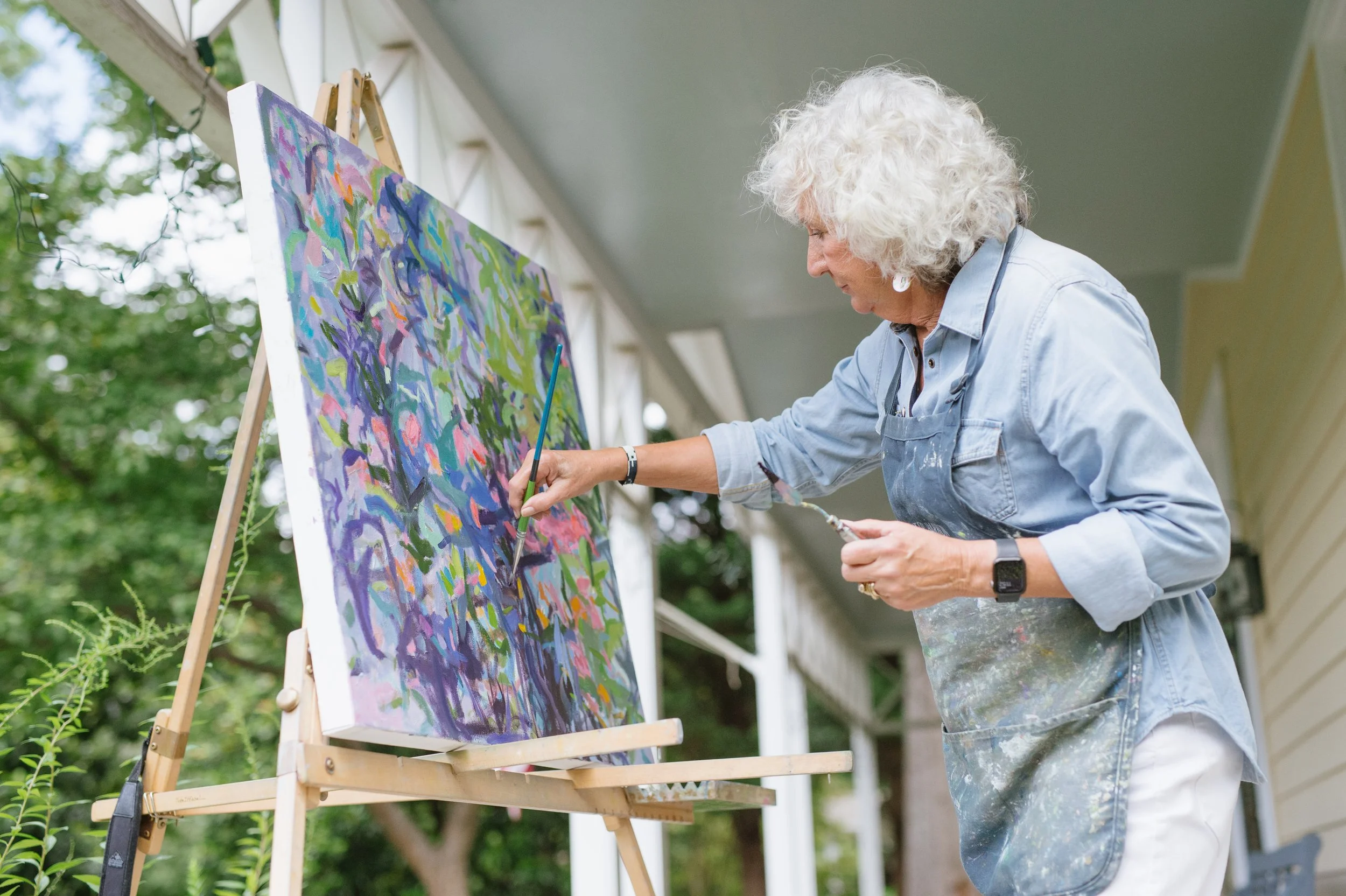 Priscilla painting en plein aire, outdoors on a colorful canvas set on an easel. She is wearing a light blue shirt and paint-splattered pants.