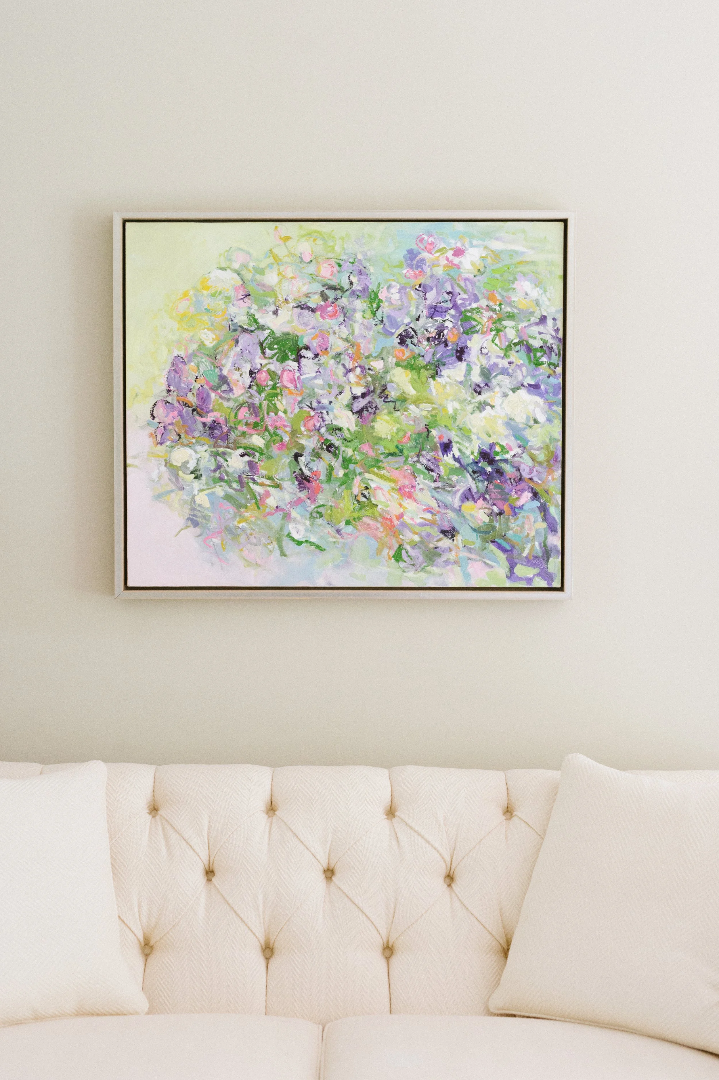 A framed abstract floral painting hangs on a light-colored wall above a white tufted sofa with matching pillows.