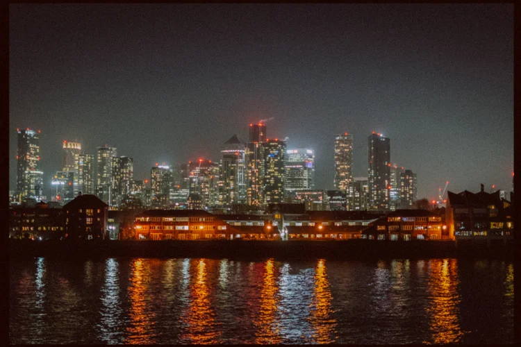 Night cityscape photograph of London's skyline across the Thames, with illuminated high-rise buildings and warm amber-lit Victorian warehouses in the foreground reflected in the dark water.