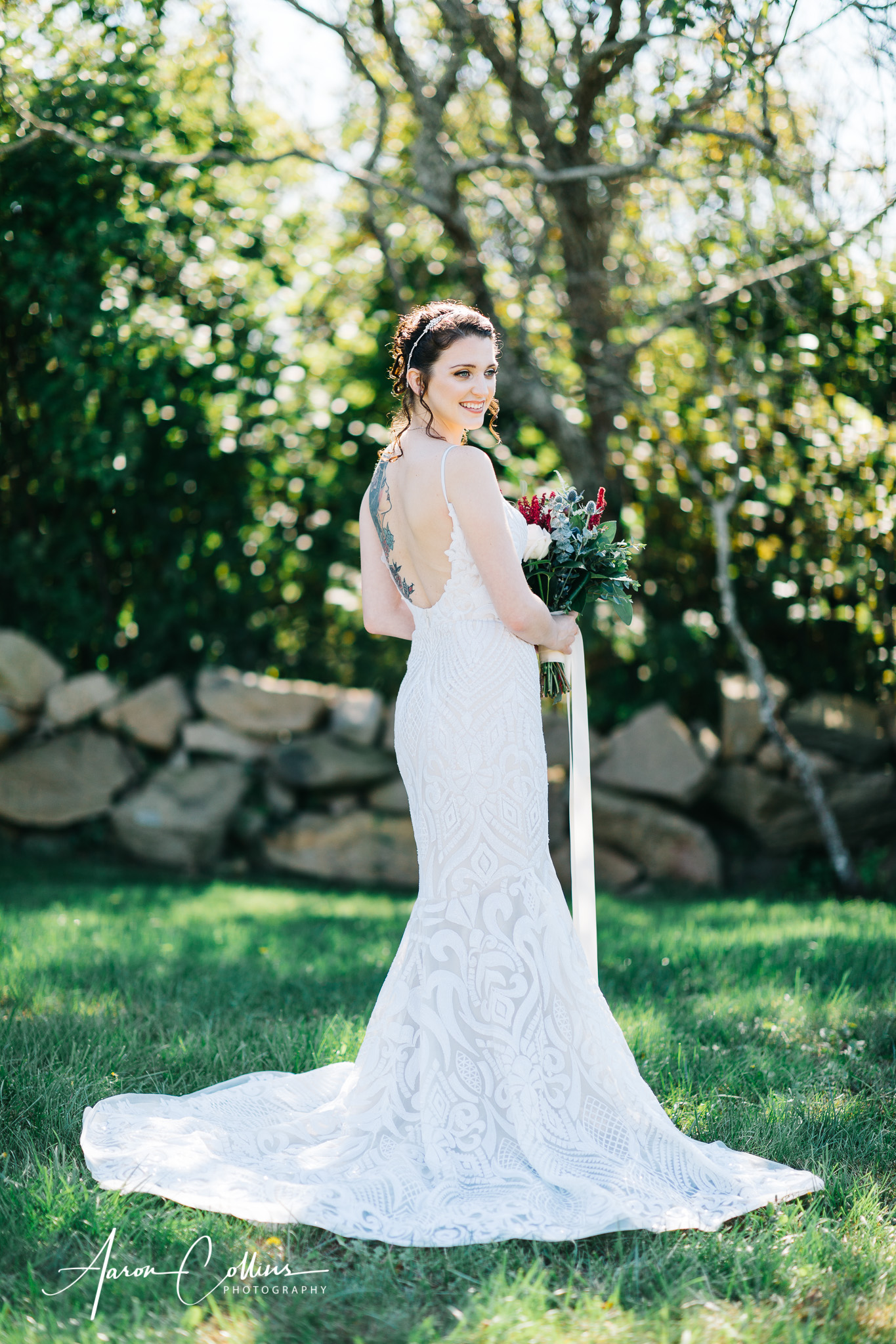 Full length portrait of the bride in her wedding dress outside in front of green leaves and grass, with her tattoo showing on the back