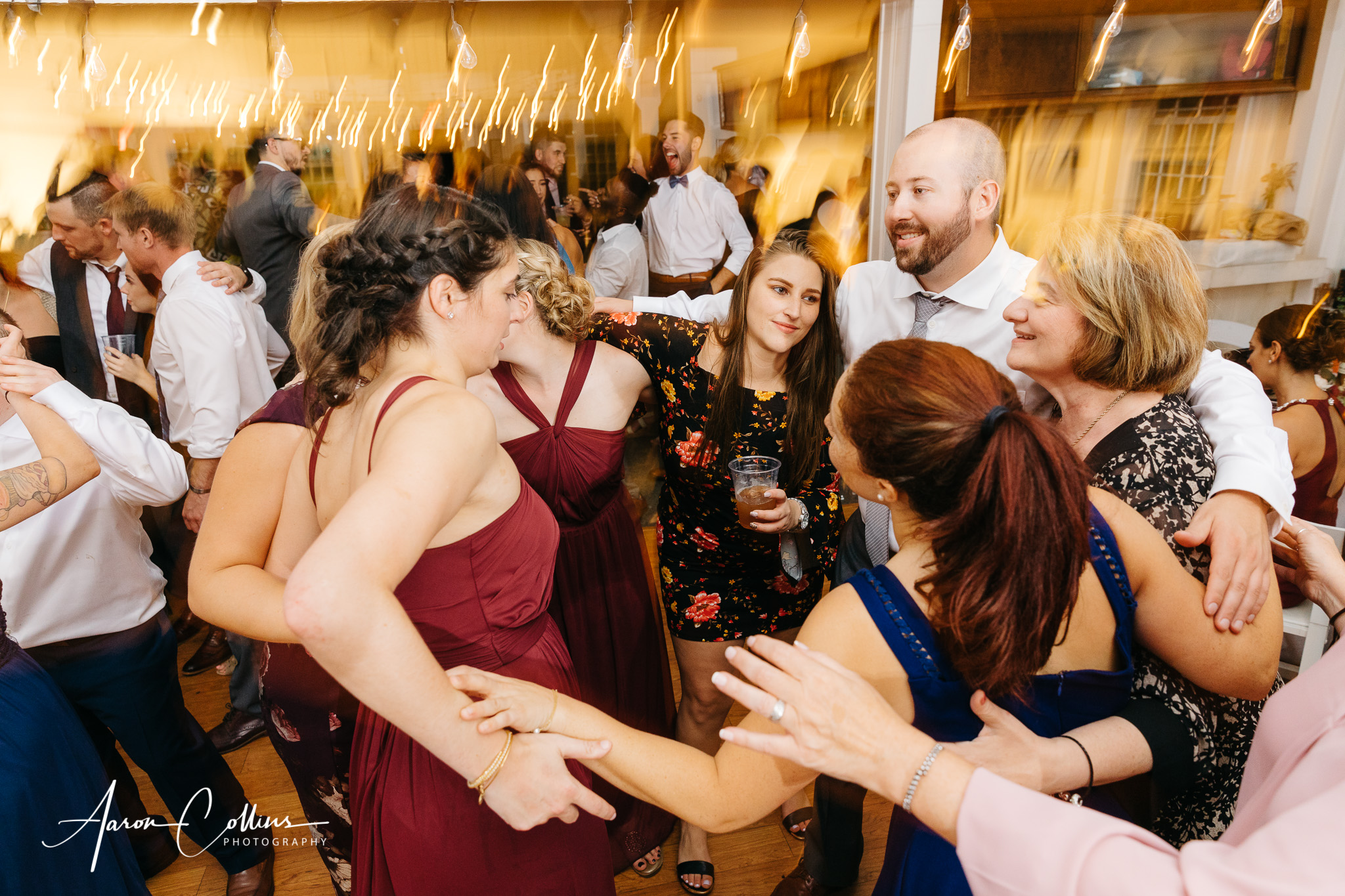 The wedding reception rages on as the night continues, with blurred lights in the background while people dance smiling. Taken with slow shutter speed for effect