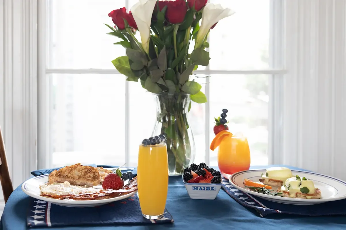 A breakfast spread on a blue tablecloth includes two plates with eggs, breakfast meats, and garnishes, two tropical drinks with fruit and berries, a bowl of mixed berries, and a vase with red and white roses in front of a window.