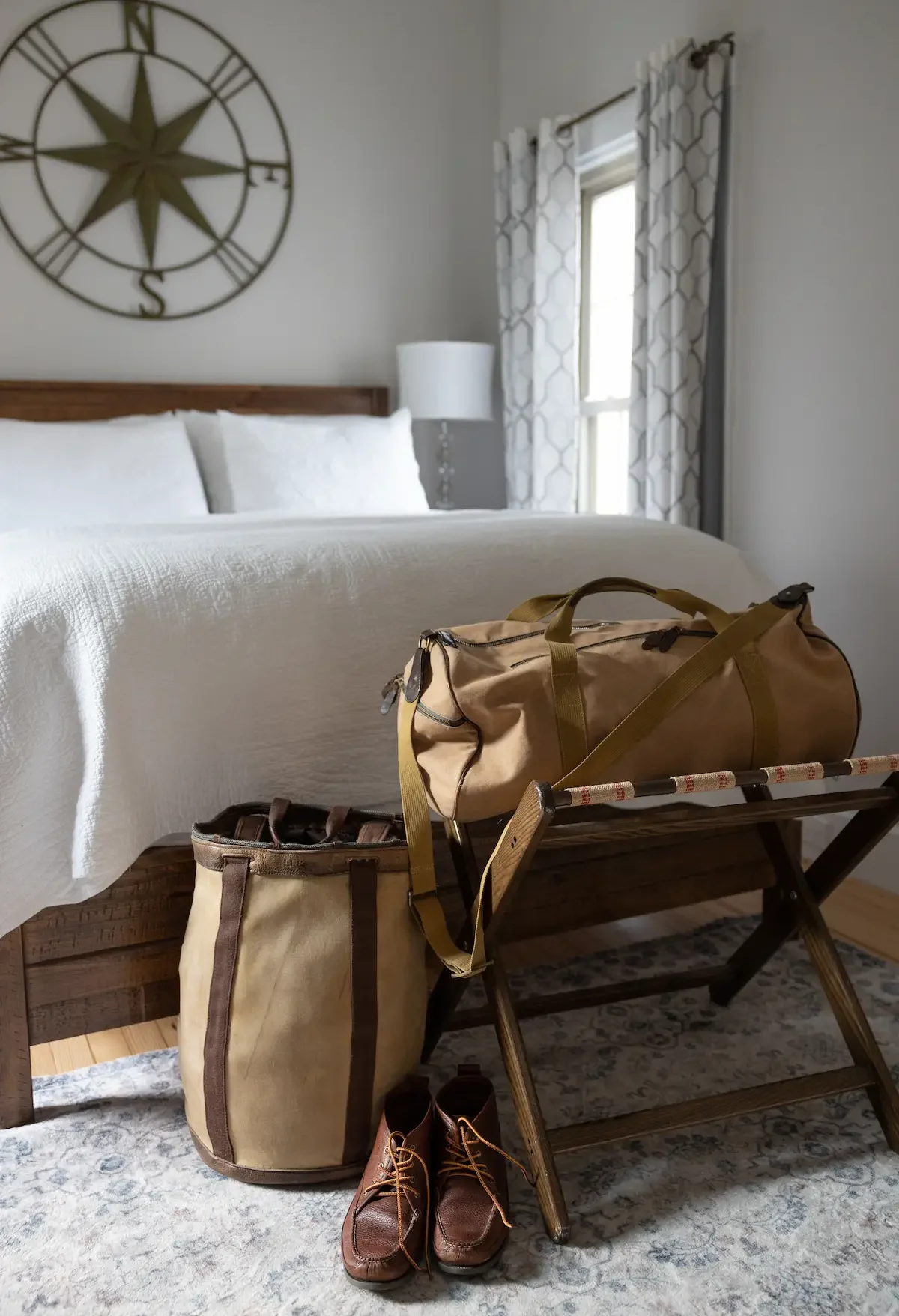 A bedroom with a bed, a beige duffel bag, a brown and beige canvas bag, a pair of brown shoes, a wooden luggage rack, a nightstand with a white lamp, and a window with patterned curtains. A large wall clock is above the bed.