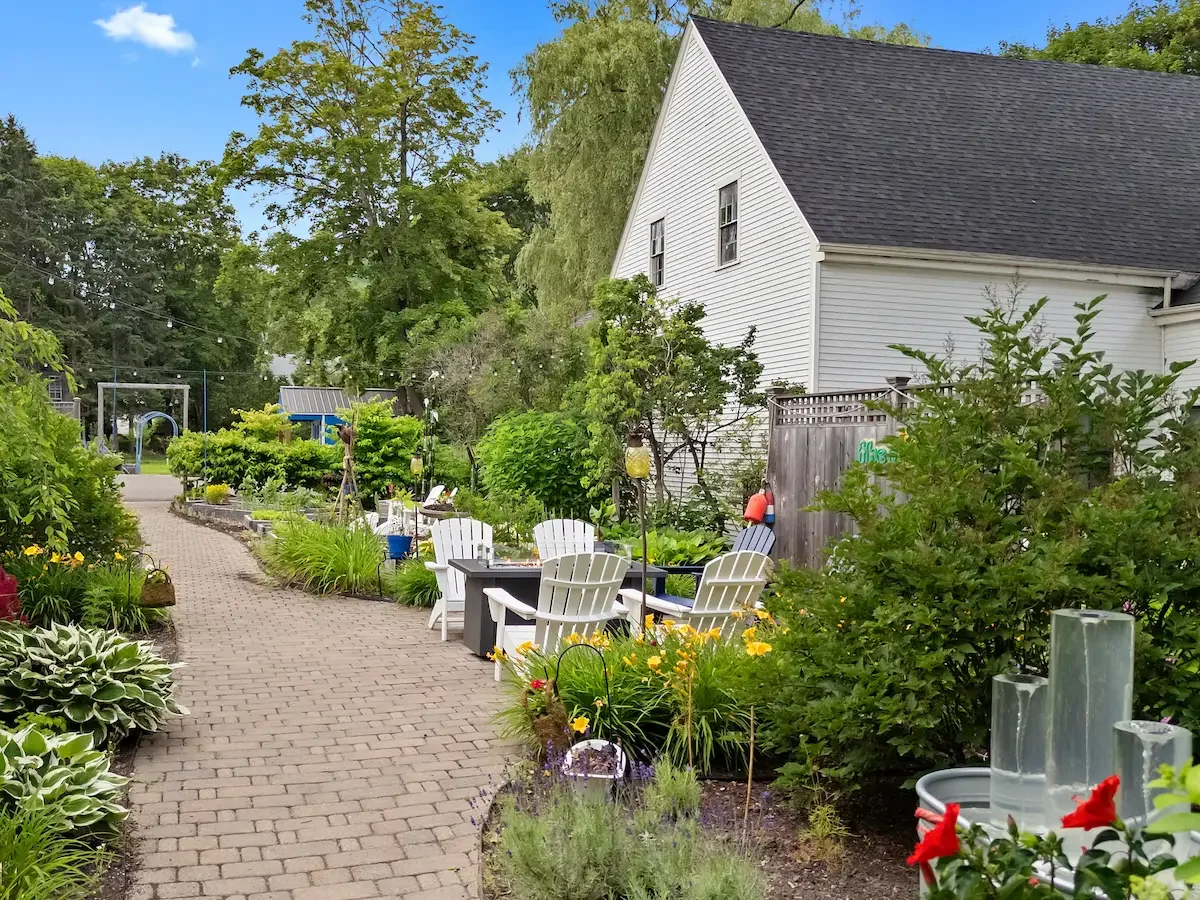 A garden with a brick pathway, white Adirondack chairs, and lush green plants and flowers next to a white house with dark roof shingles.