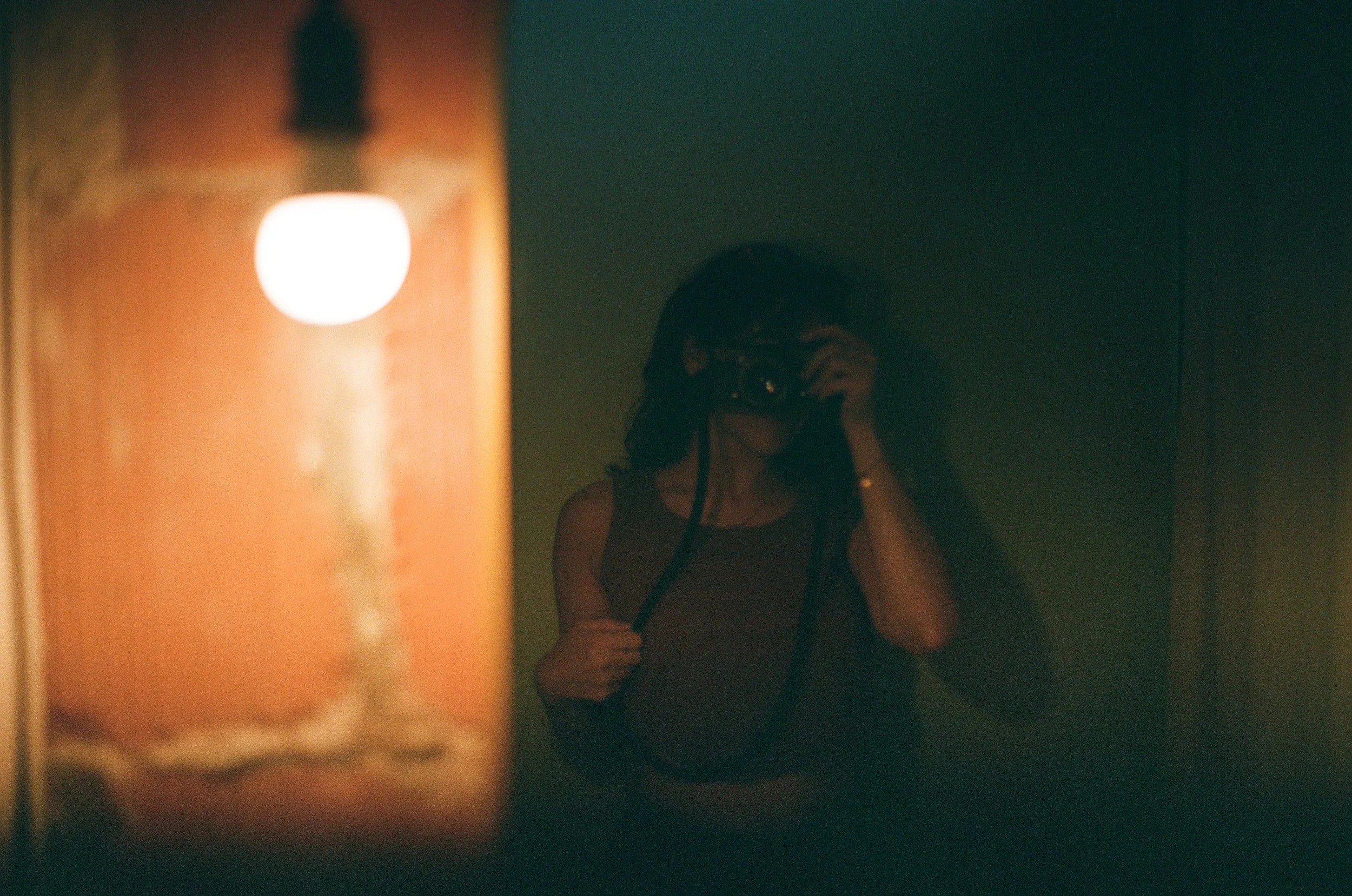 A woman taking a photo of her reflection in a dark mirror, with a blurred orange lamp in the foreground.