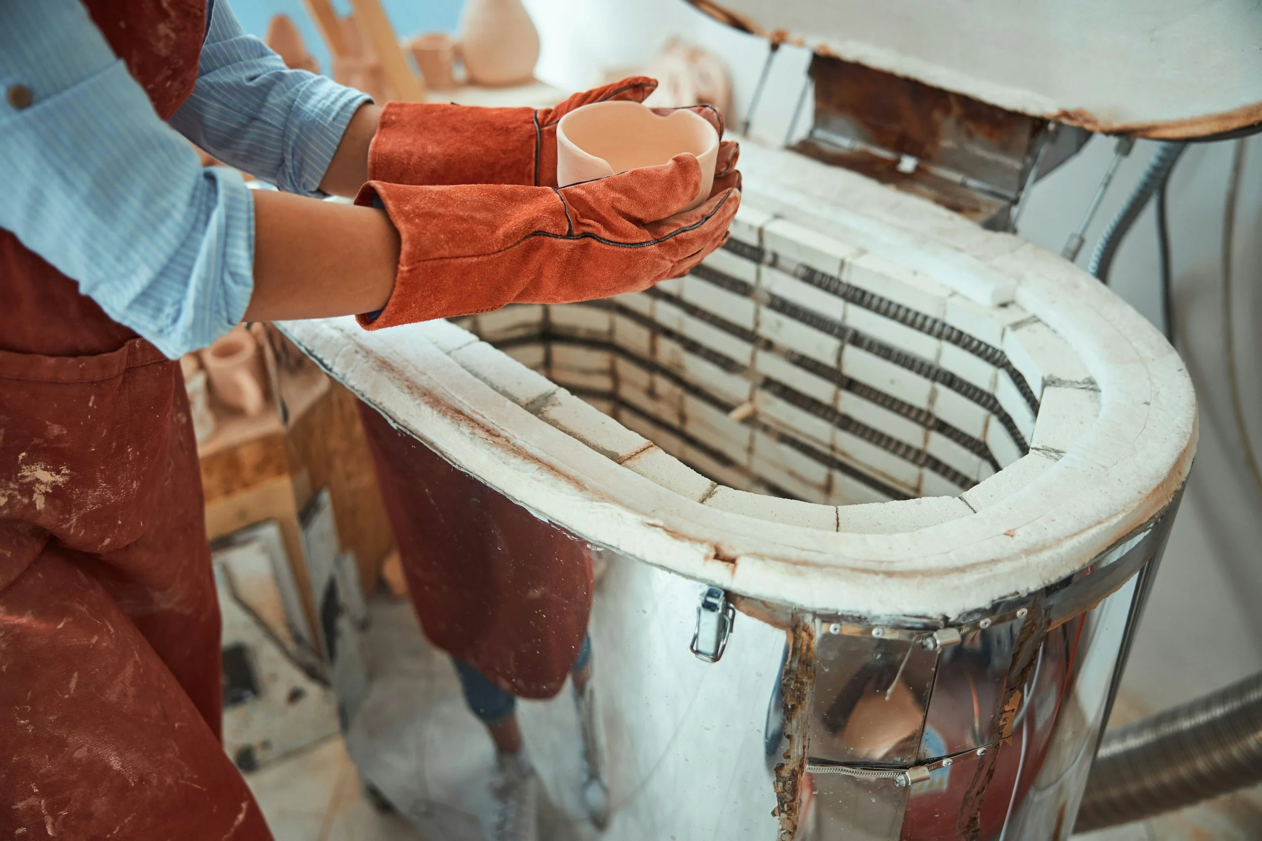 Person wearing gloves and an apron handling a ceramic piece over a kiln in a workshop.