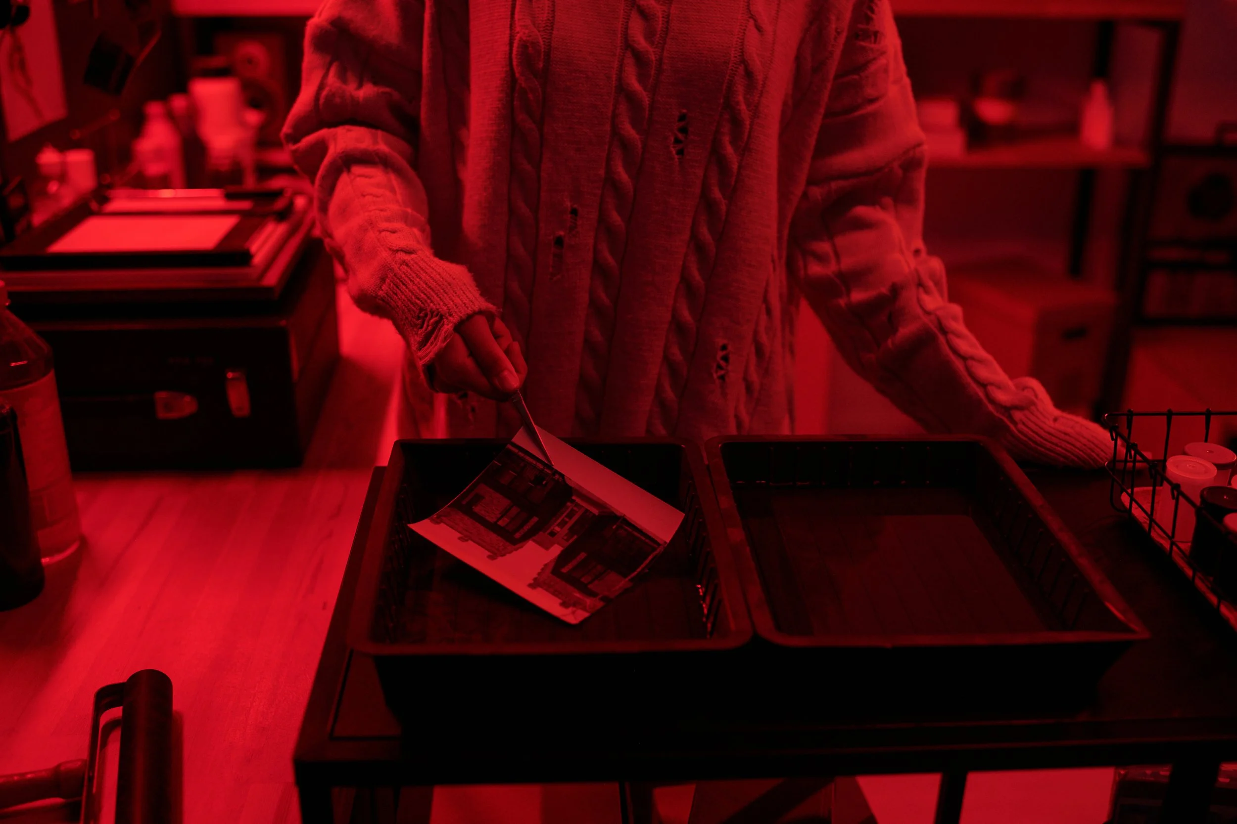 Black and white print being developed by hand in darkroom trays under red safelight.