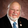 A portrait of a smiling older man with glasses, a white beard, and white hair, wearing a black suit, white shirt, and blue tie, with pins on his suit lapel against a mottled brown background.