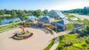 Aerial view of a greenhouse complex with solar panels on roofs, a circular paved area with a colorful sculpture fountain, landscaped garden, and a river with trees in the background.