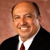 Close-up portrait of a smiling man with short, graying hair, wearing a dark suit, white shirt, and red tie, against a brown mottled background.