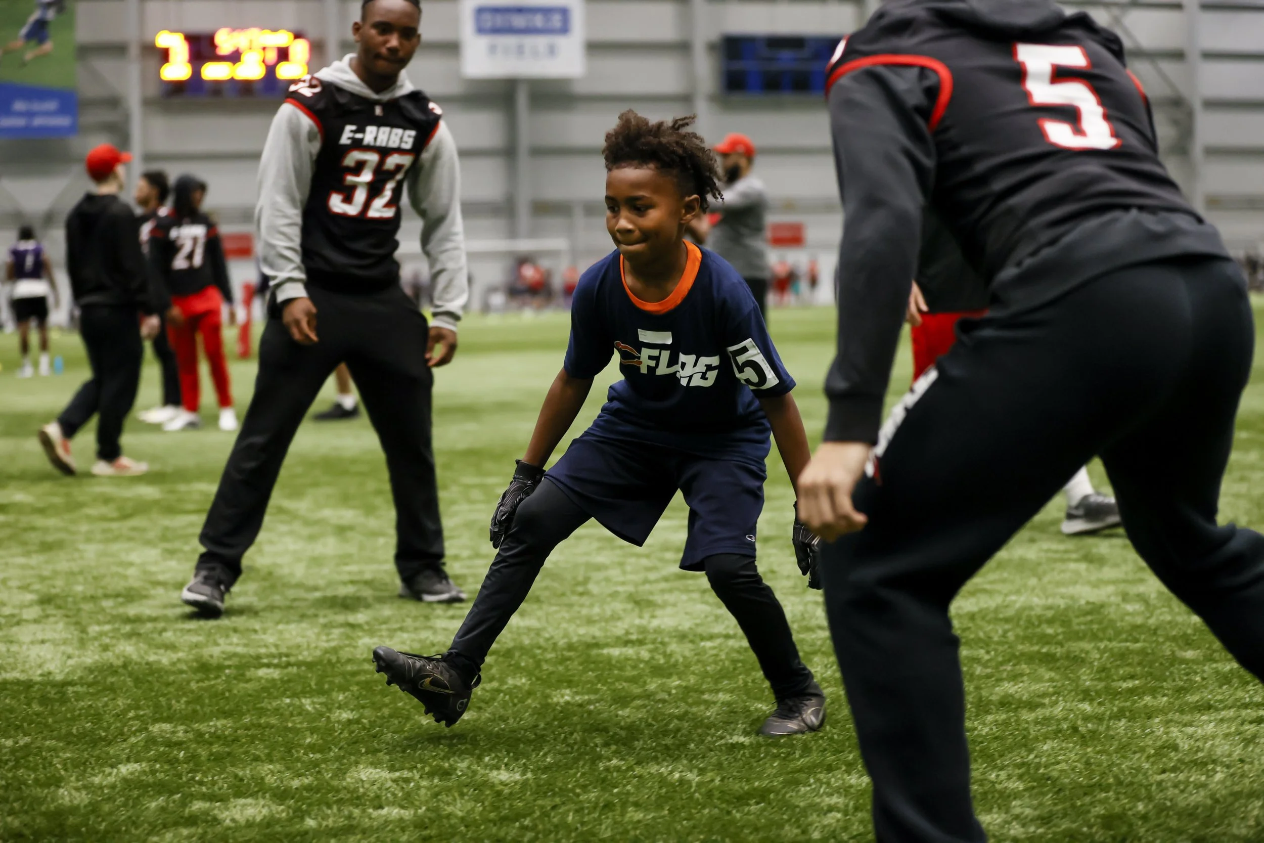 A young boy wearing a blue football uniform and black gloves is playing football on an indoor field, with two adult men in black and red sports gear around him, and other players and spectators in the background.