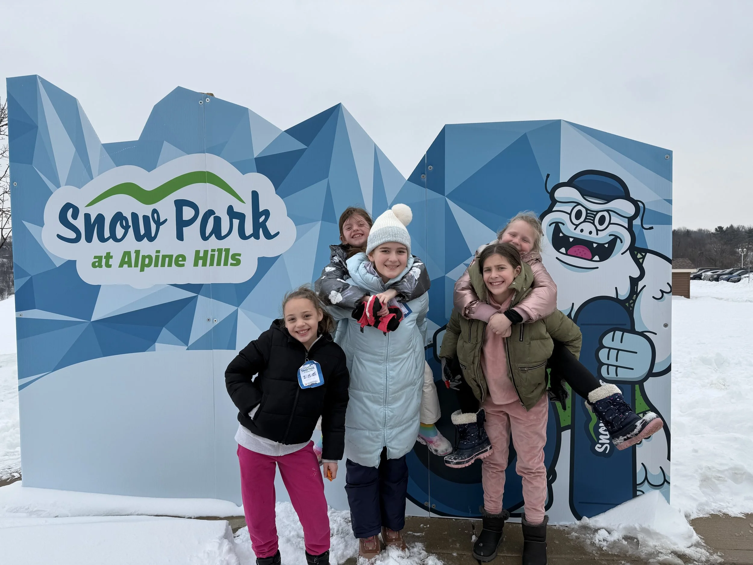 Four children in winter clothing smiling and posing at Snow Park at Alpine Hills, with a blue geometric mountain backdrop and a cartoon yeti giving a thumbs-up.
