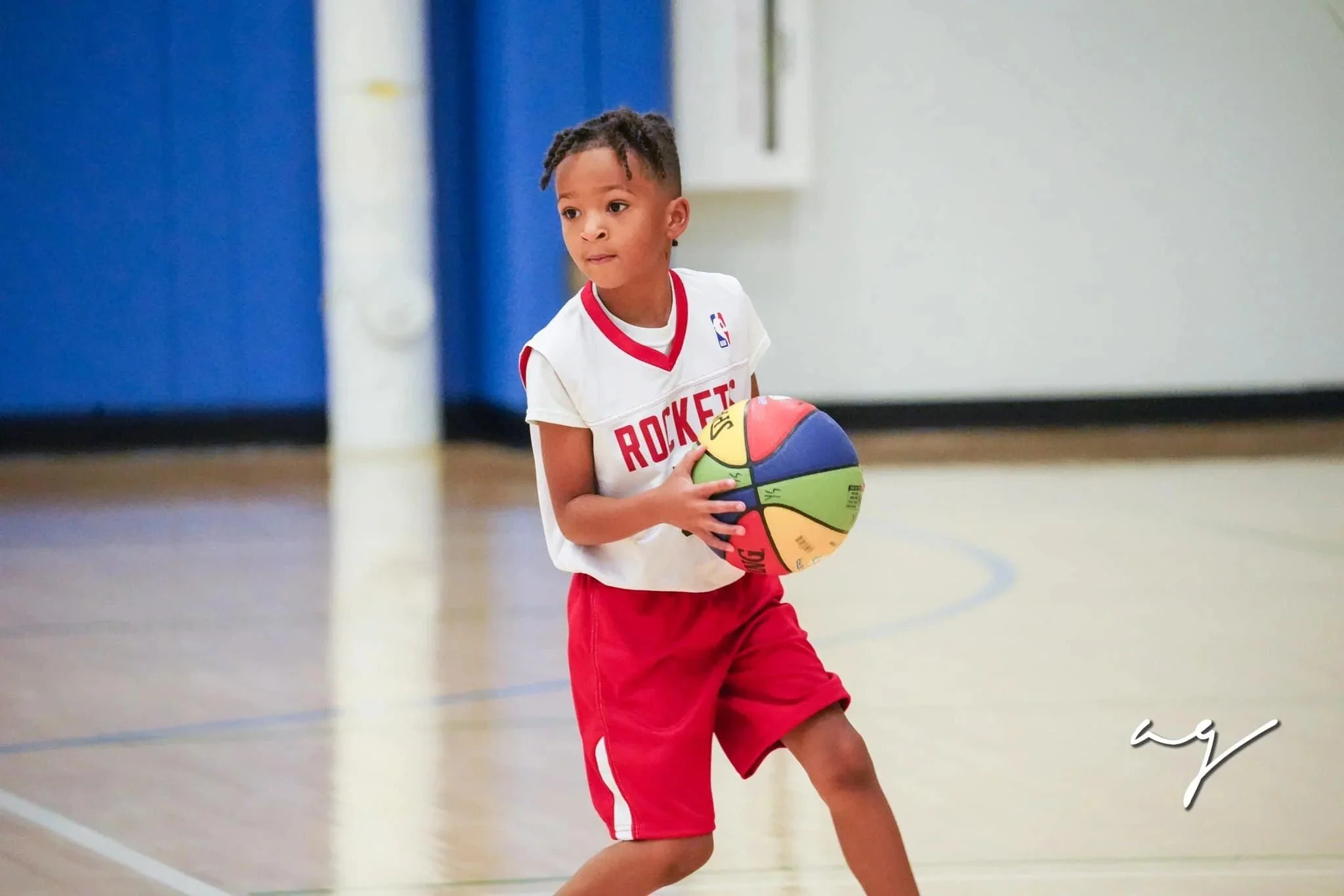 A young boy wearing glasses and a black basketball jersey with "KINGS" written on it, running on an indoor basketball court during a game or practice, with other children and adults in the background.