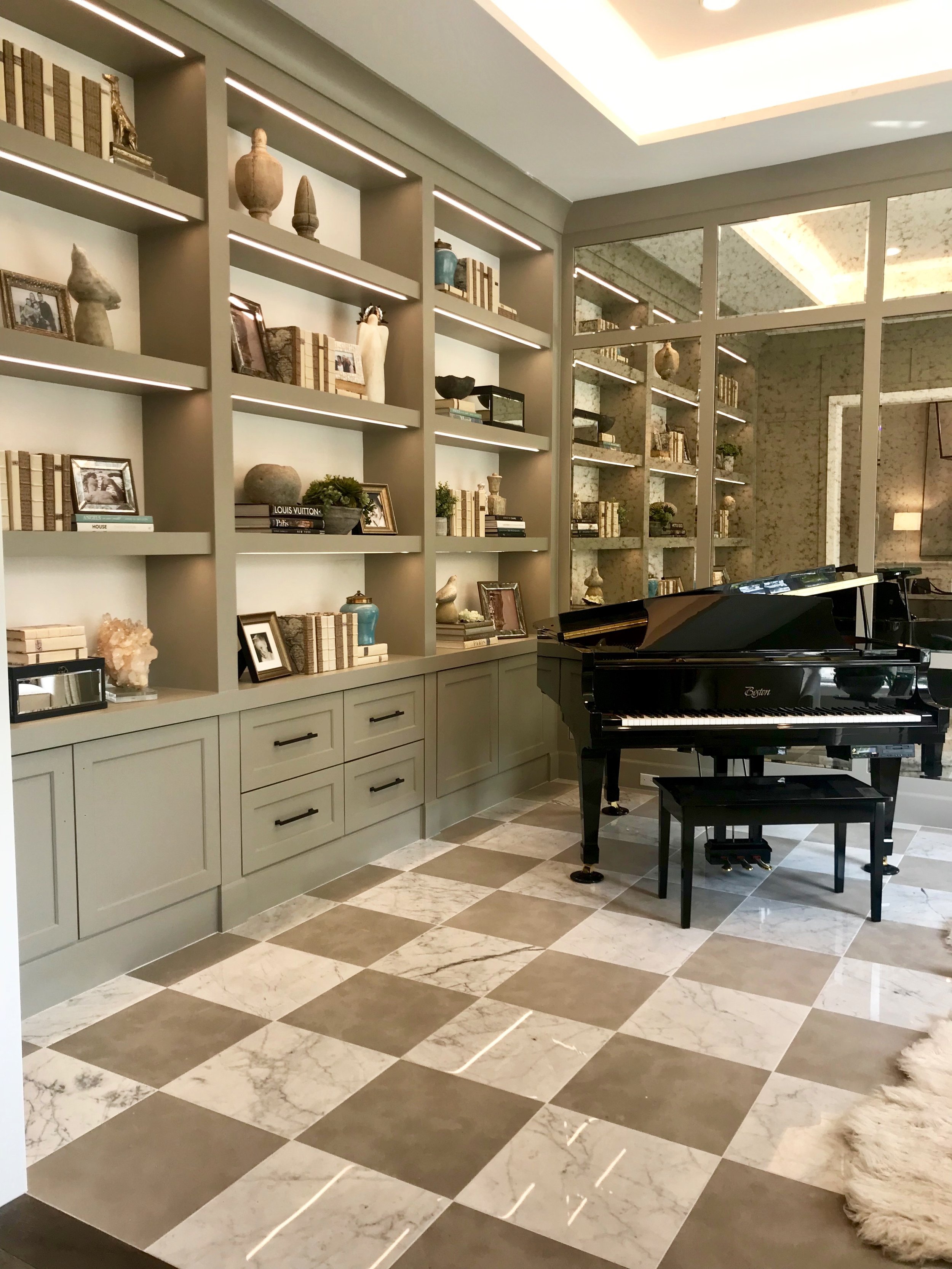 Warm gray wood paneling surrounds the library with beveled antique mirror insets at the focal wall, reflecting elegance at all angles, and a glossy black Boston baby grand piano welcomes guests from the adjacent entry.