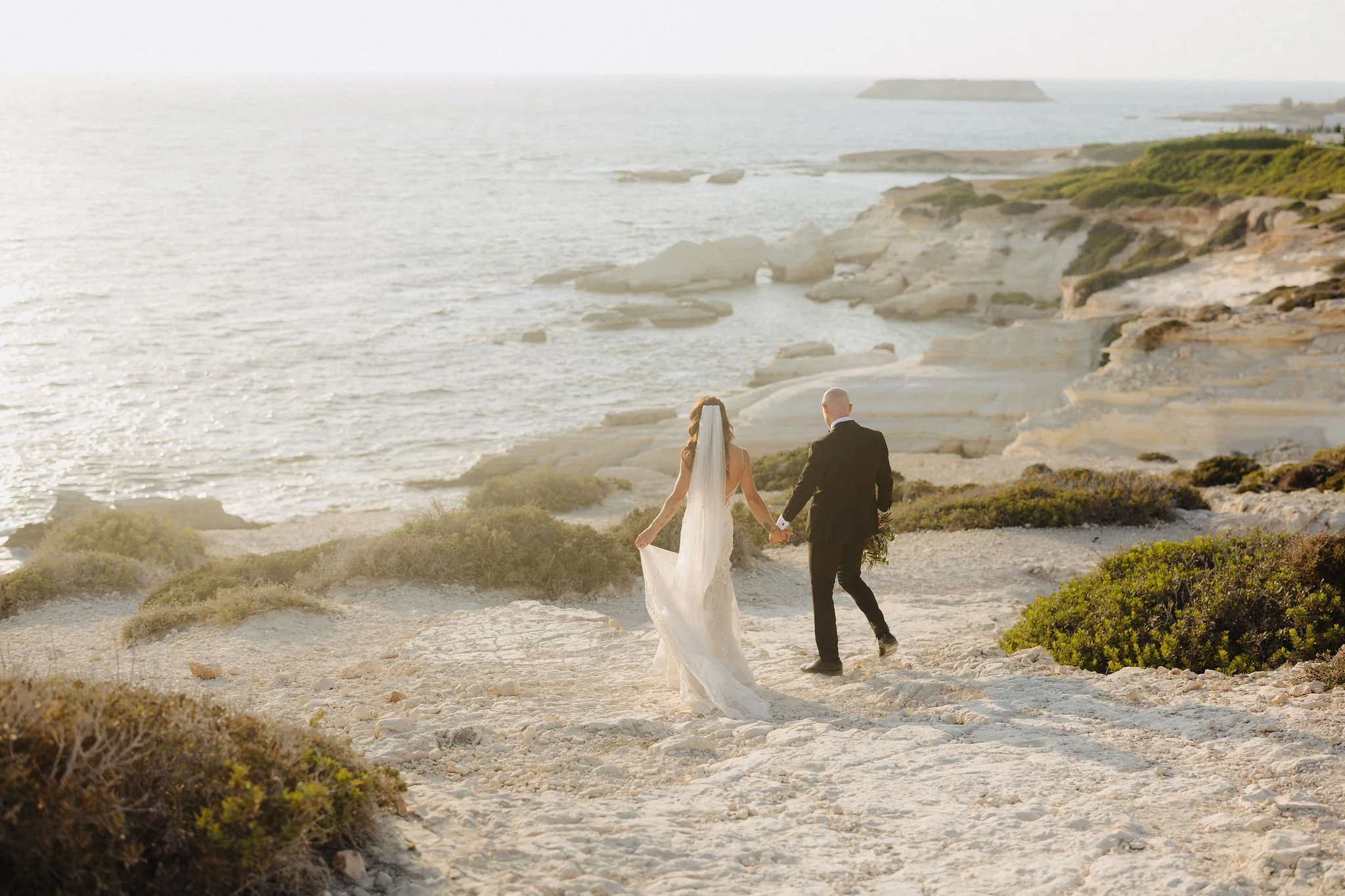 A bride and groom walking hand in hand along a rocky beach with cliffs and the ocean in the background, during a sunset or sunrise.