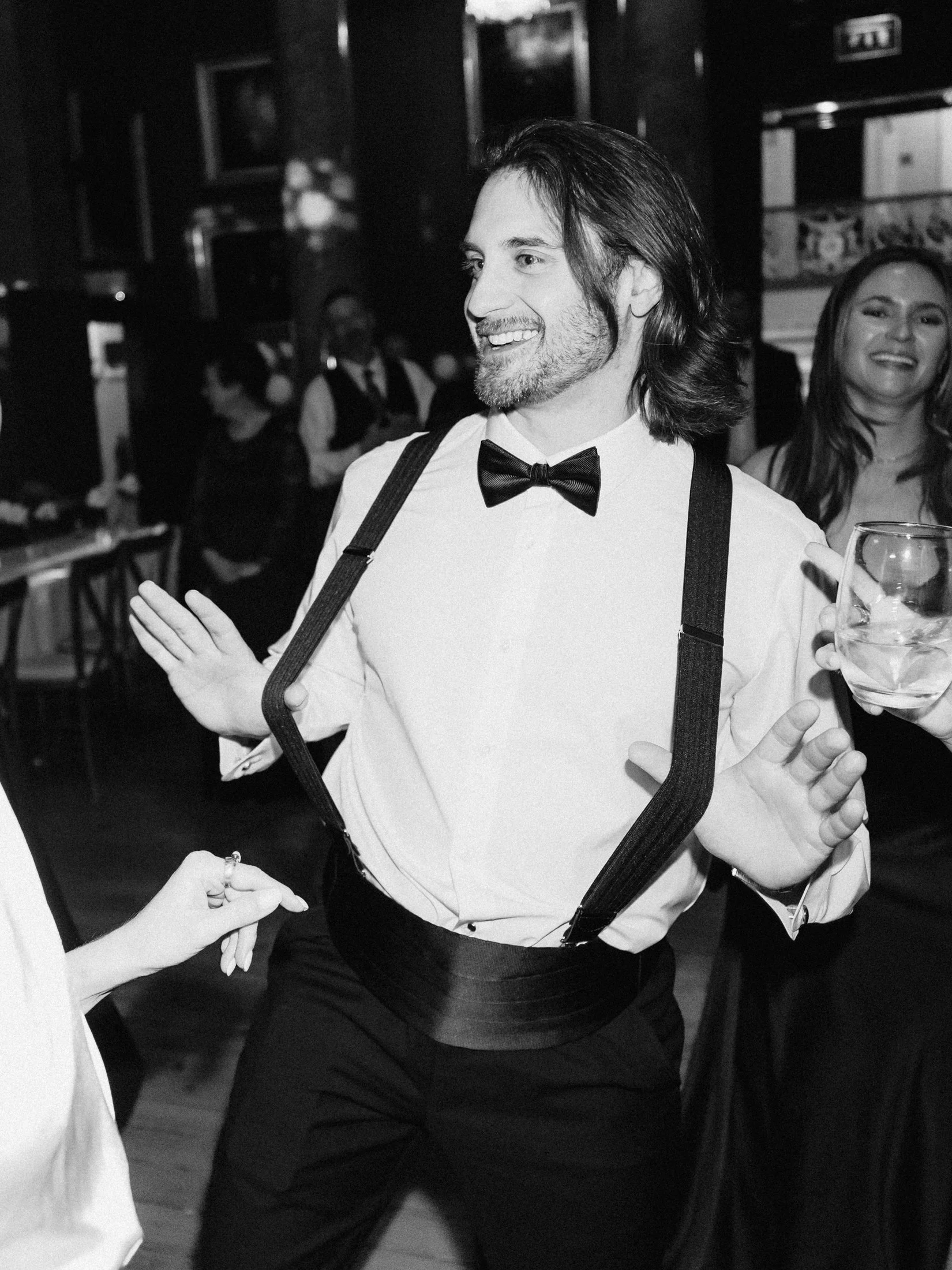 A black-and-white photo of a man with long hair, beard, and mustache, dressed in a white shirt, black bow tie, suspenders, and black pants at a social event. He is smiling and holding a clear glass of drink in one hand. People are visible in the back